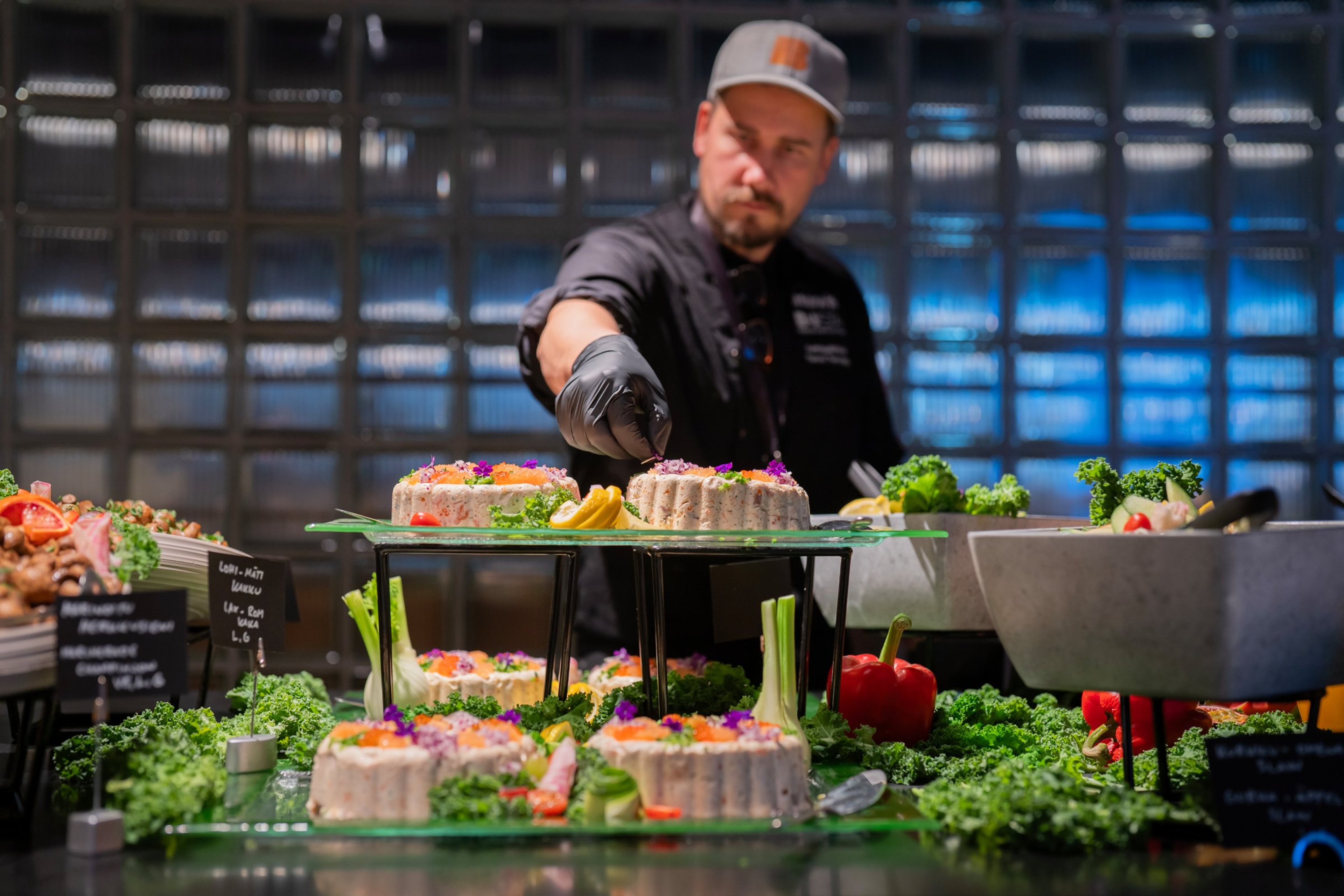 Kokki mustissa käsineissä asettelee koristeellisia voileipäkakkuja ja muita ruokia tarjoilupöydälle. Taustalla näkyy sininen lasiseinä.
