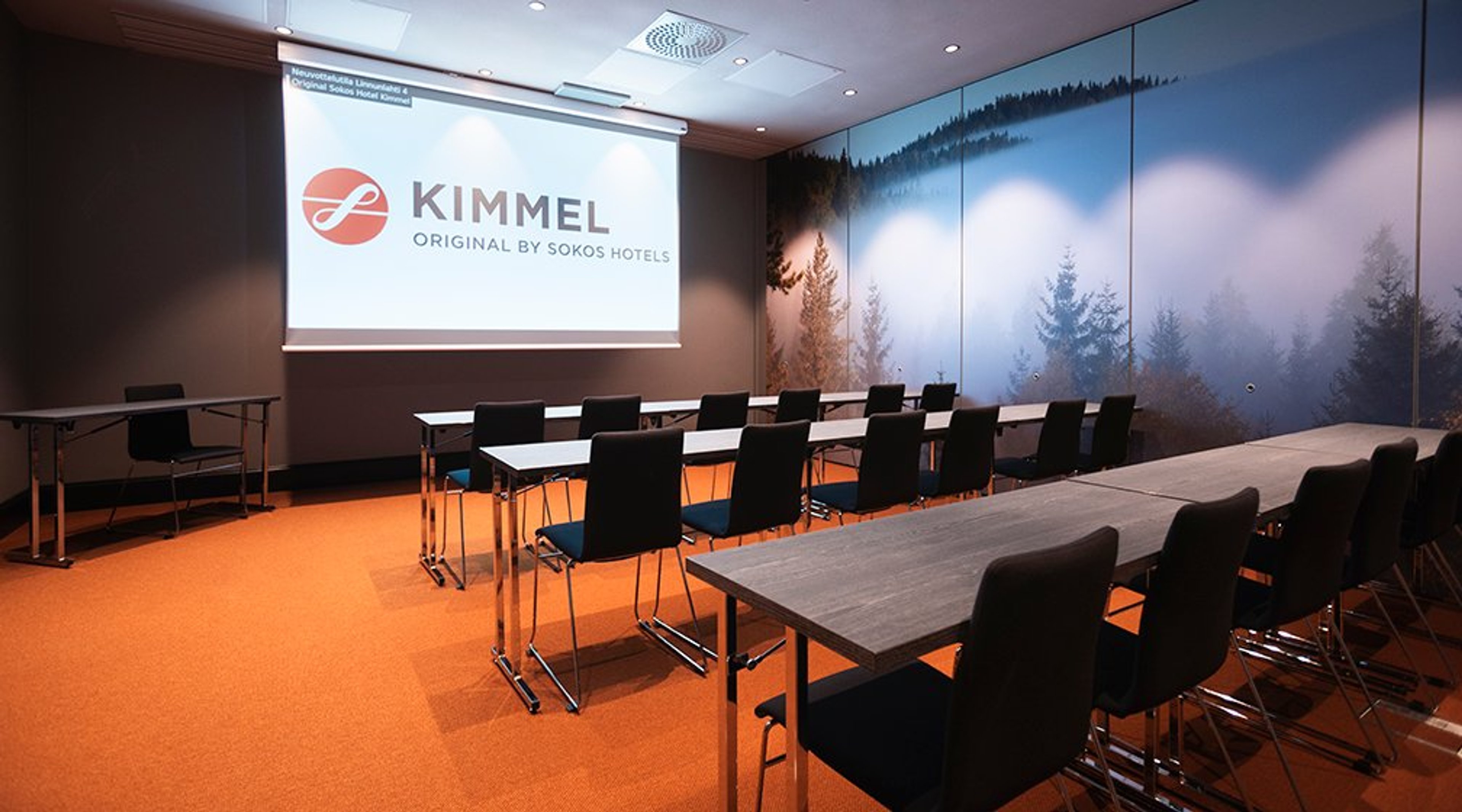 Kokoushuone oransilla lattialla, valkoisia pöytiä mustine tuoleineen ja metsäaiheinen seinämaalaus. Valkokankaalla Kimmel-logo.