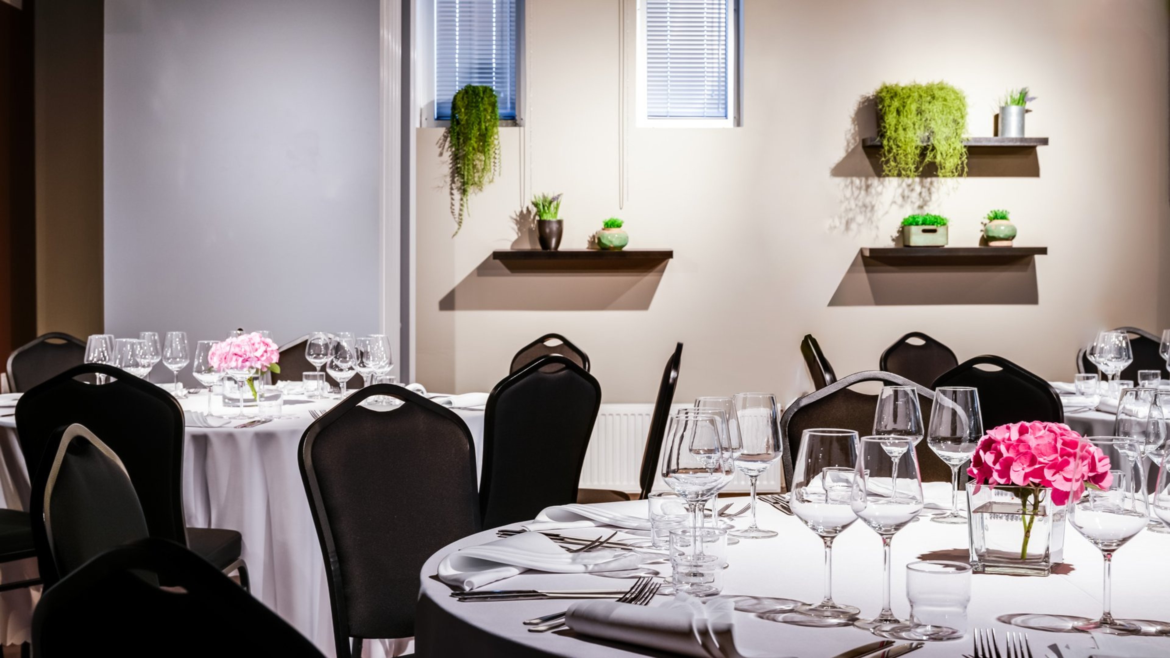 Juhlatila, jossa on valkoisten pöytäliinojen peittämiä pyöreitä pöytiä, mustia tuoleja ja viinilaseja. Seinällä viherkasvisommitelmia hyllyillä, pöydillä pinkkejä kukkia maljakoissa.