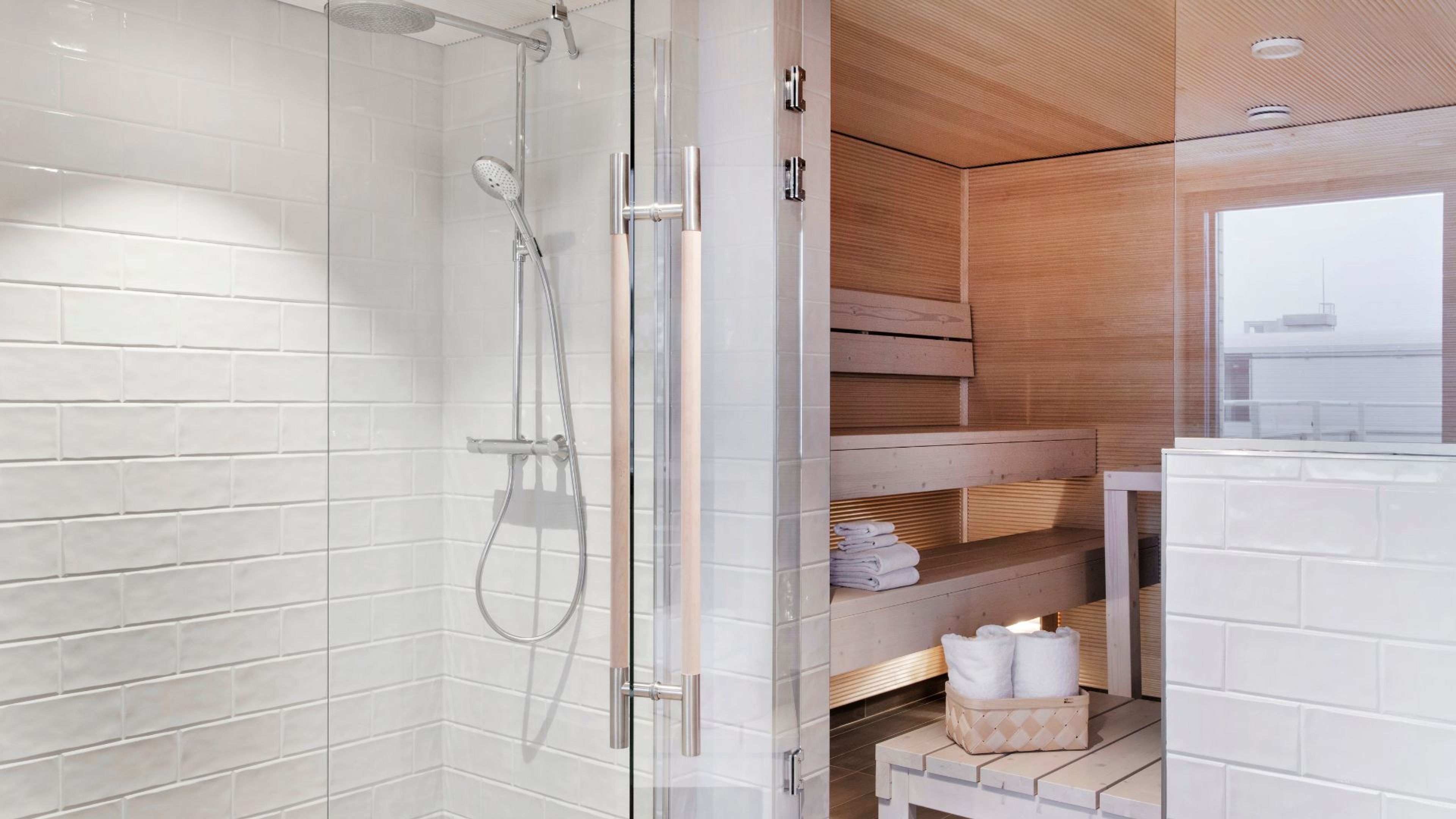 Moderni kylpyhuone, jossa lasinen suihkukaappi ja viereinen puupaneloitu sauna valkoisilla pyyhkeillä.