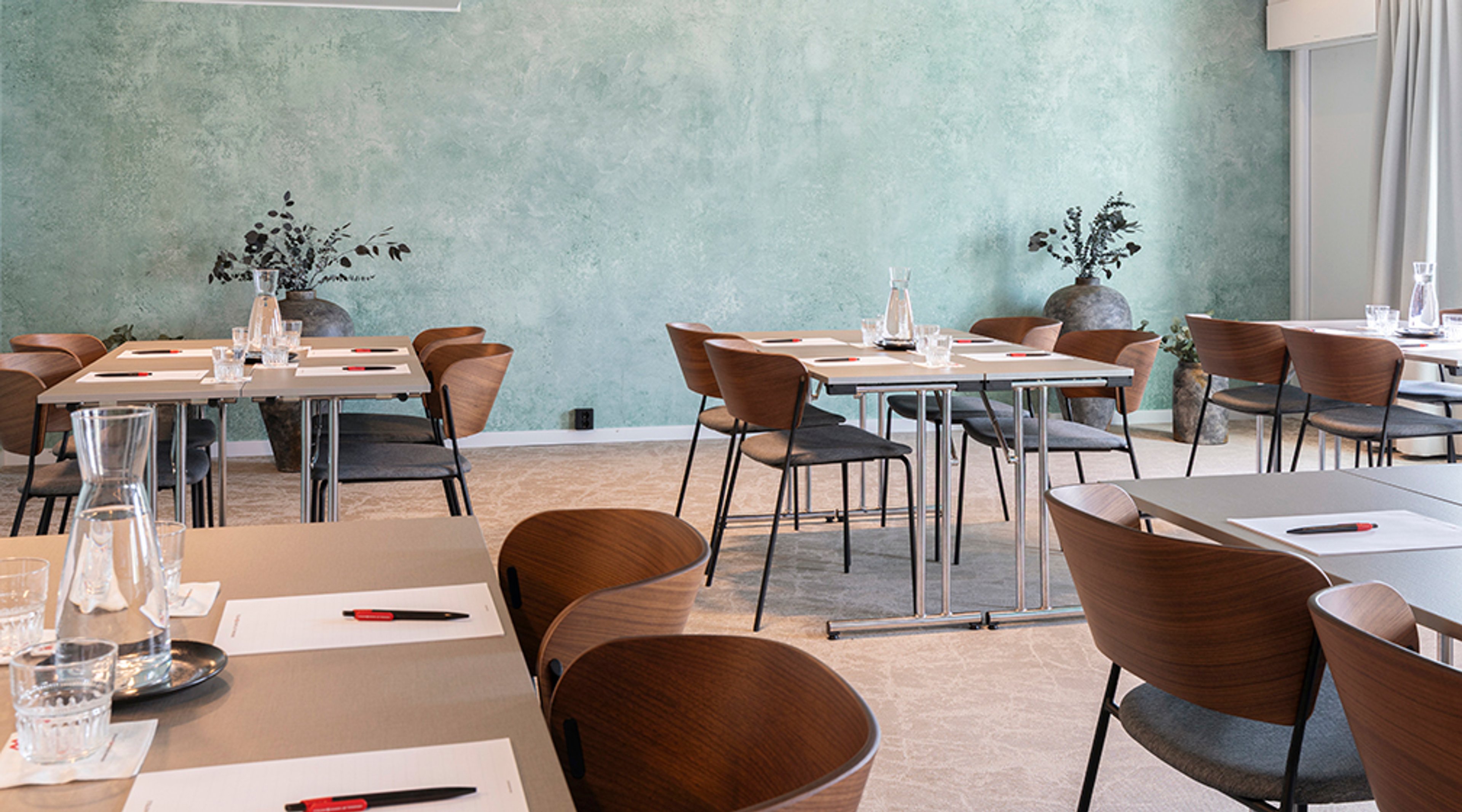 Moderni kokoustila, jossa vaaleita pöytiä ja puisia tuoleja. Turkoosi tekstuuripintainen seinä ja pieniä viherkasveja pöydillä. Tasainen lattia.