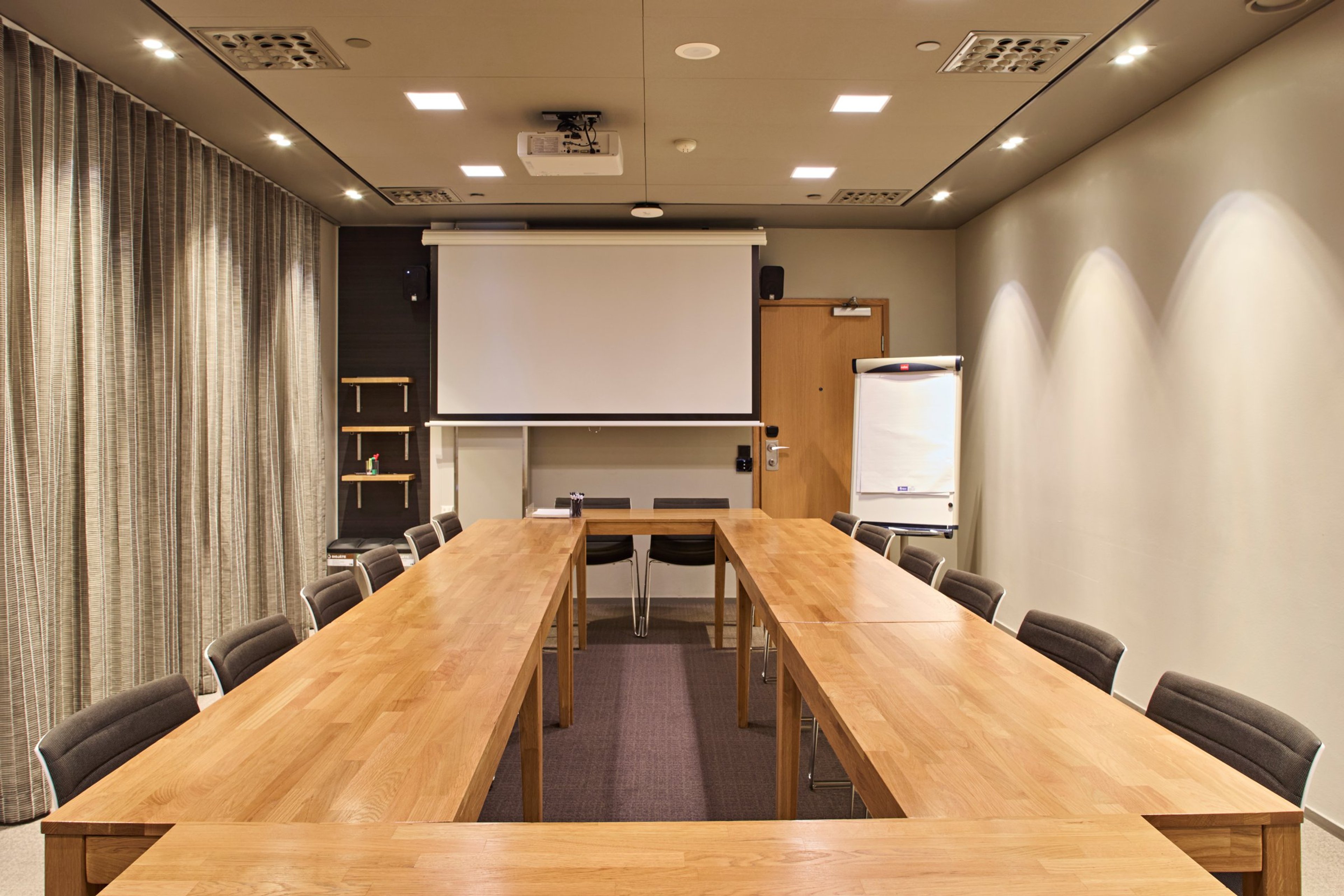 Kokoushuone puisilla pöydillä, tummilla tuoleilla, valkoisella projektorikankaalla ja fläppitaululla. Tasainen valaistus ja kova lattiapinta.