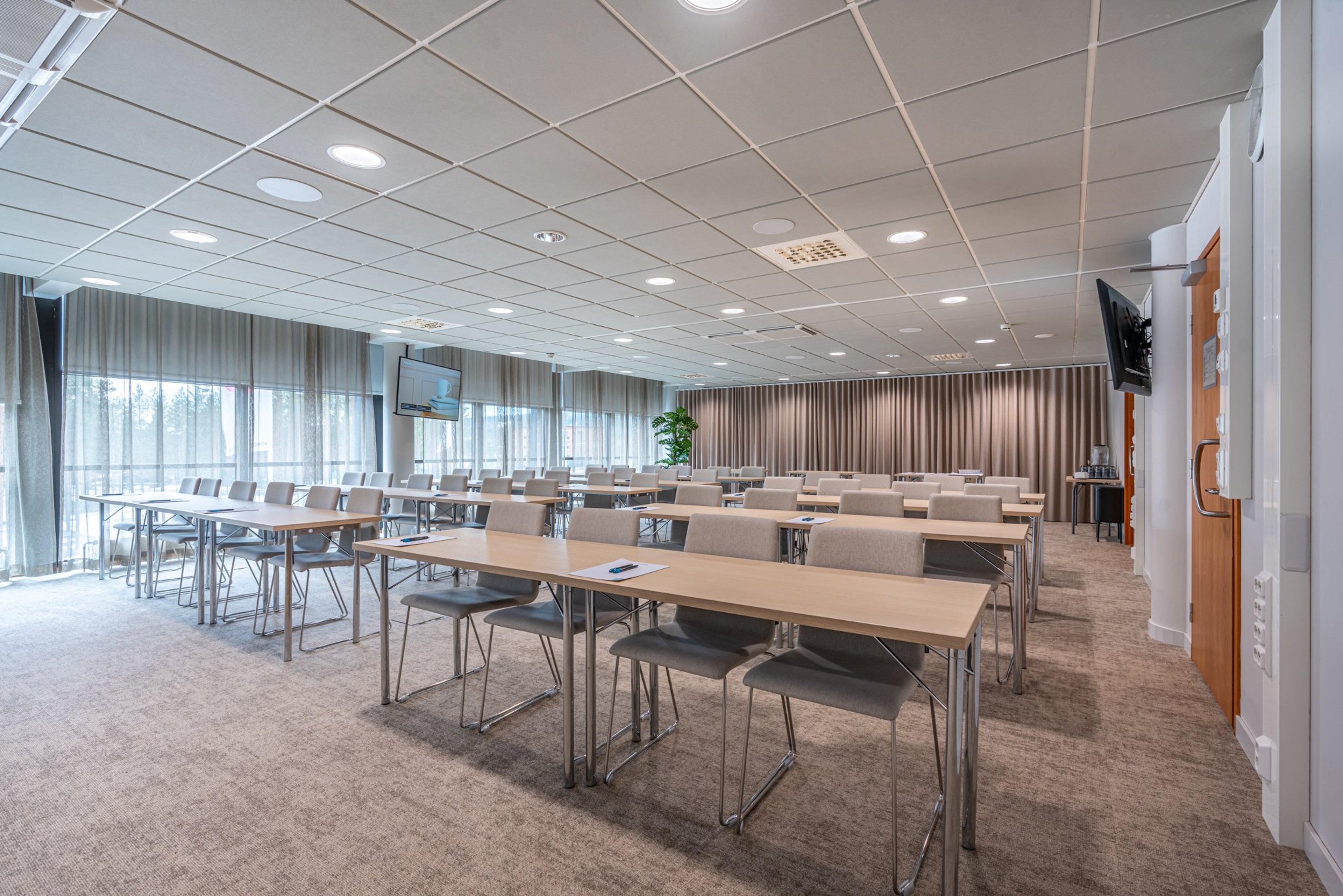 Valoisa kokoustila riveittäin järjestetyillä pöydillä, beige kokolattiamatto ja suuret ikkunat. Seinällä näyttö ja huoneen perällä verhot.