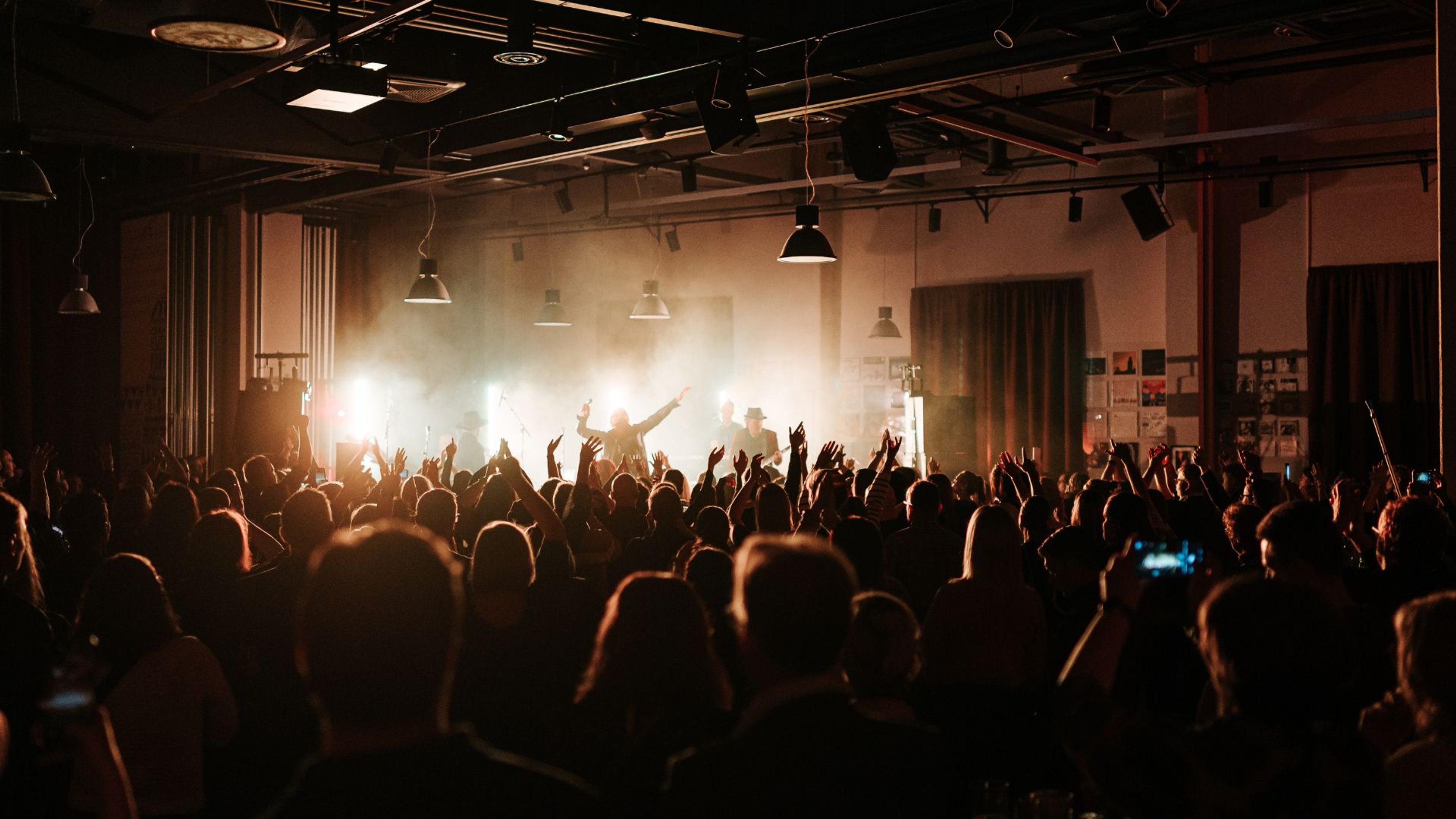 Konsertti pienessä intiimissä tilassa, jossa yleisö nostaa käsiään ilmaan esiintyjien edessä. Lavalla on kirkas valotekniikka.