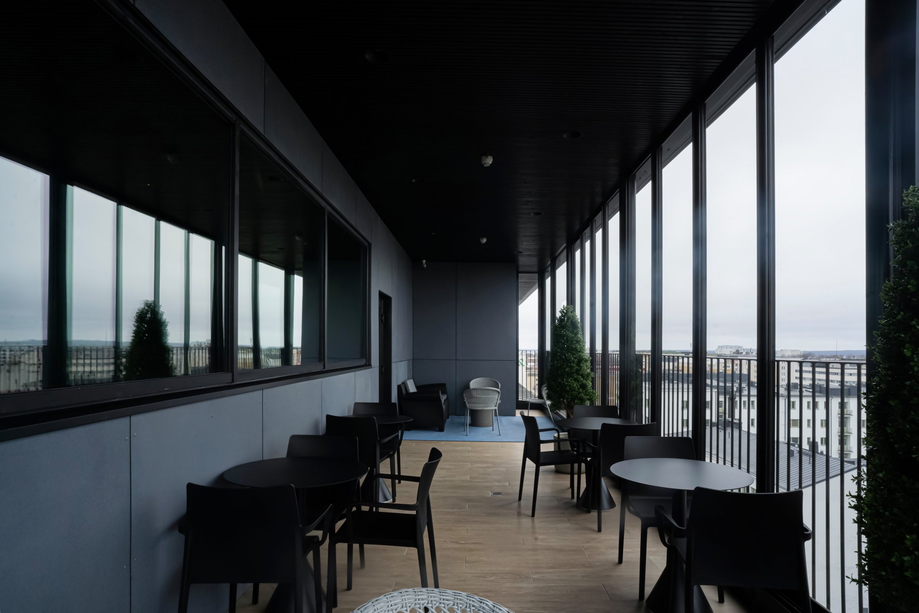 Moderni terassi korkealla rakennuksessa, jossa mustia pöytiä ja tuoleja, puulattia ja panoraamanäkymä kaupungista. Tasainen kulkuväylä.