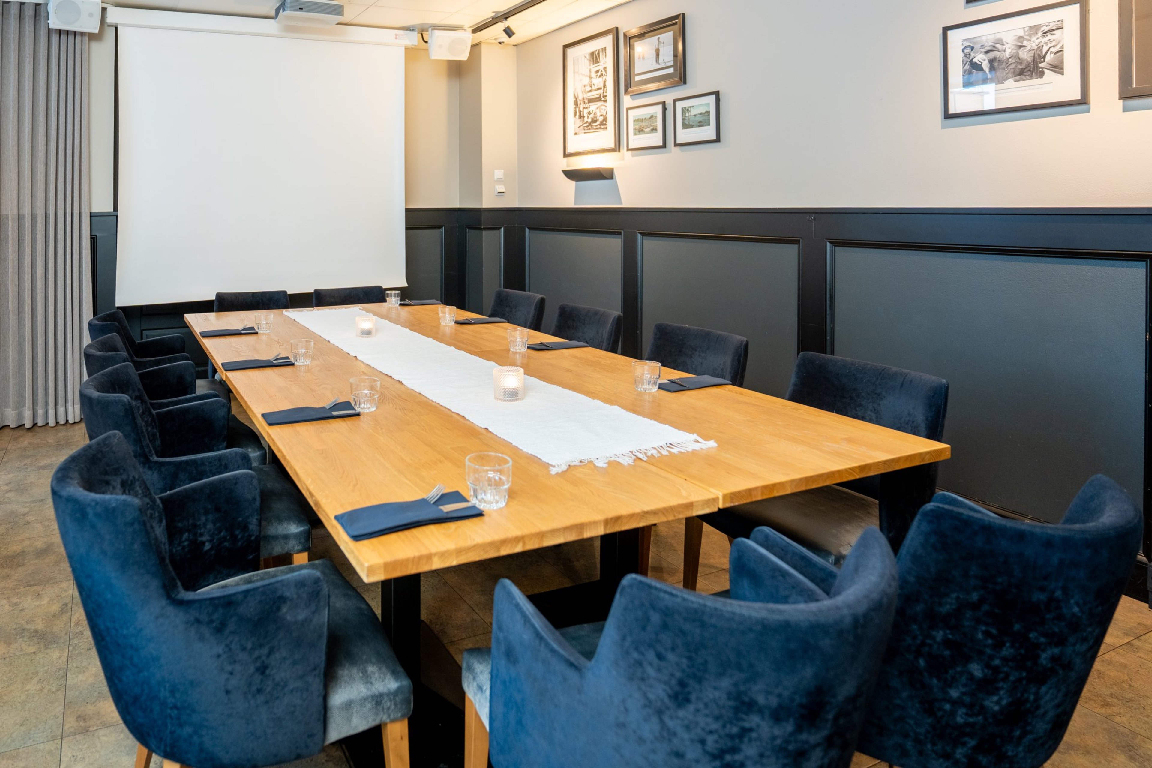 Kokoushuone, jossa puinen pöytä, siniset nojatuolit, valkoinen pöytäliina ja vesilasit. Seinillä tauluja, taustalla valkokangas.