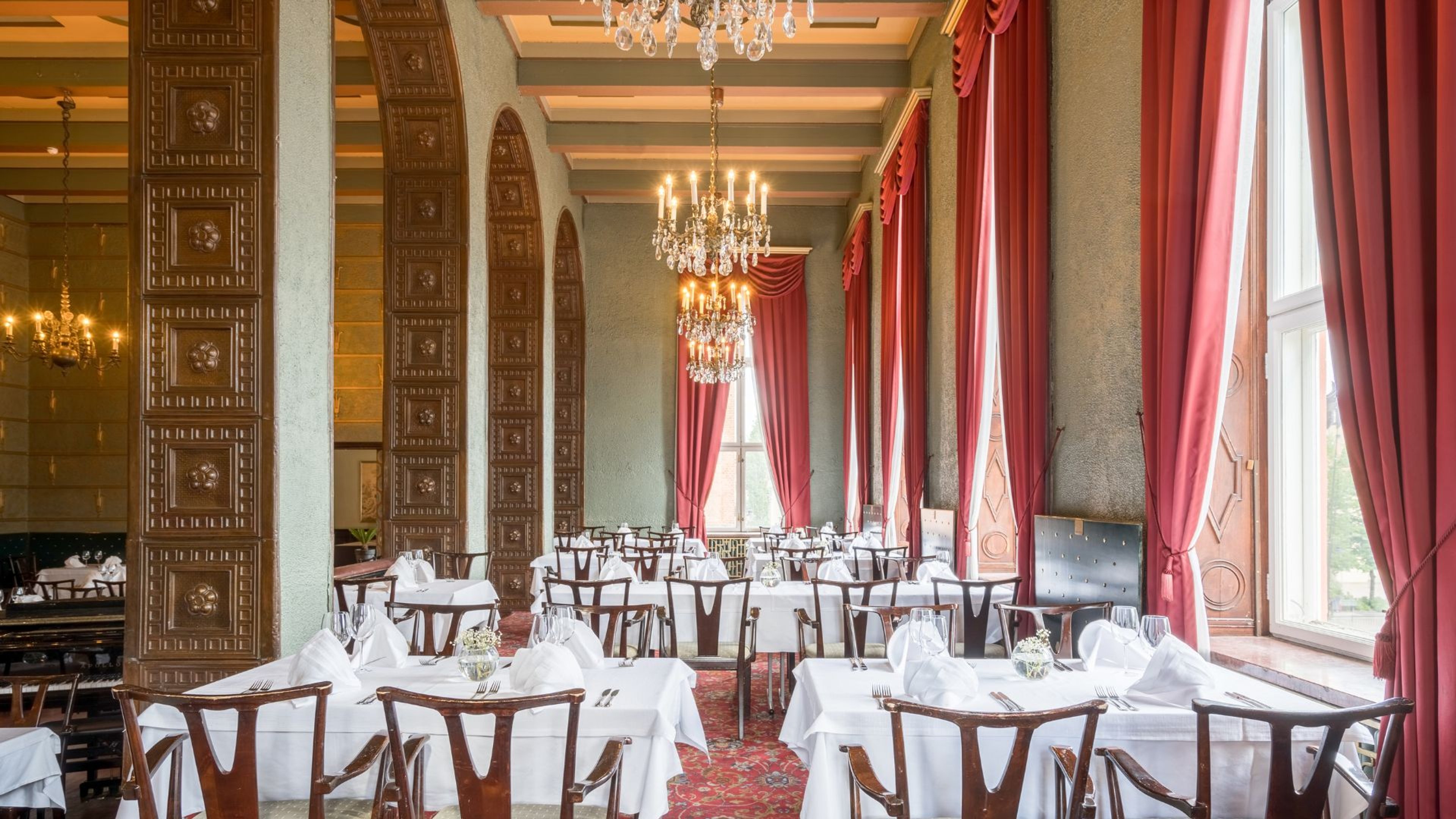 Ylellinen ravintolasali, jossa koristeelliset pilarit, kristallikruunut ja punaiset verhot. Valkoliinaiset pöydät ja puiset tuolit sijoiteltu avaraan tilaan.
