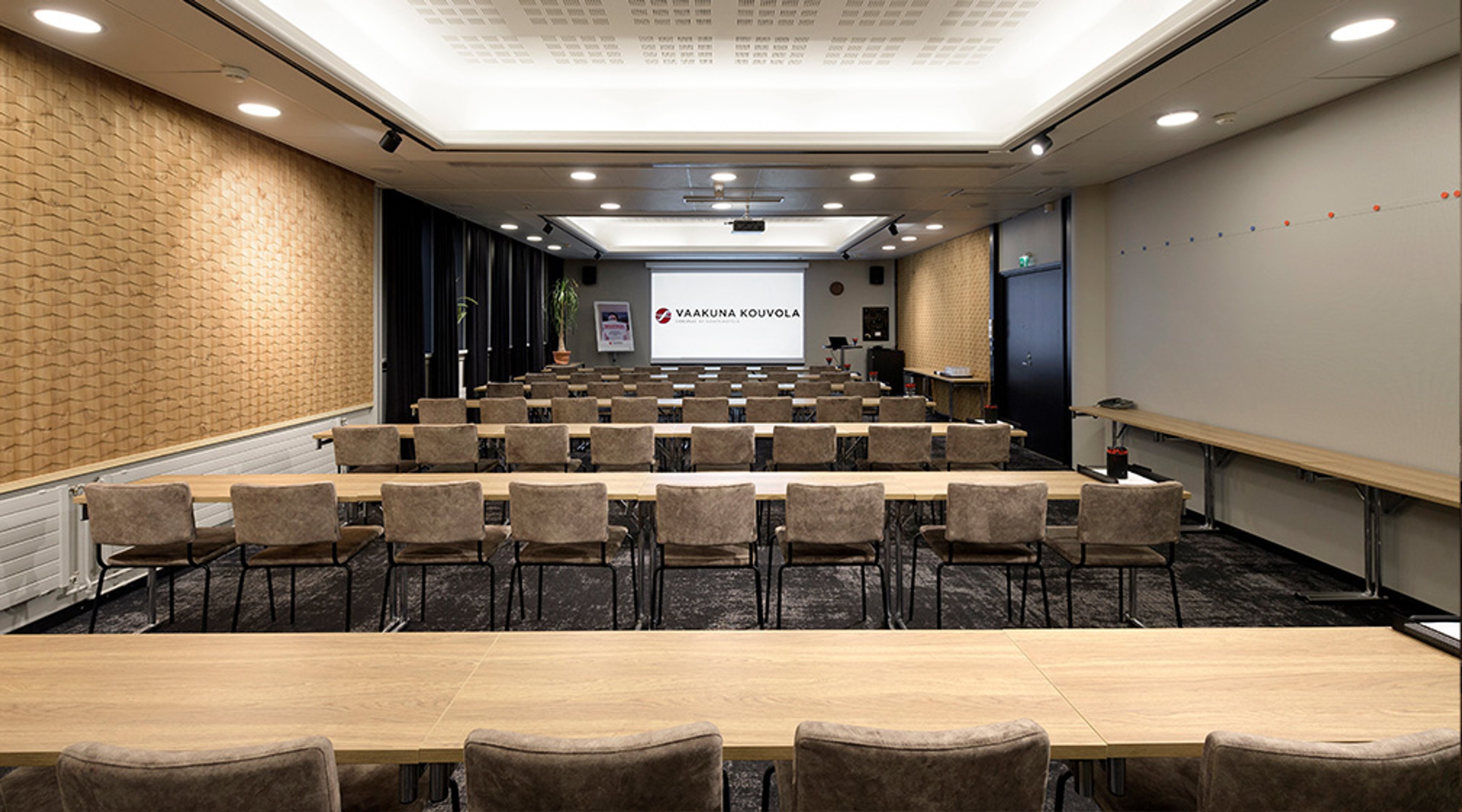 Auditoriotila jossa on riveihin asetellut pehmustetut tuolit, puinen kaarevapintainen etuseinä ja vaalea projektorikangas. Tila on hyvin valaistu.