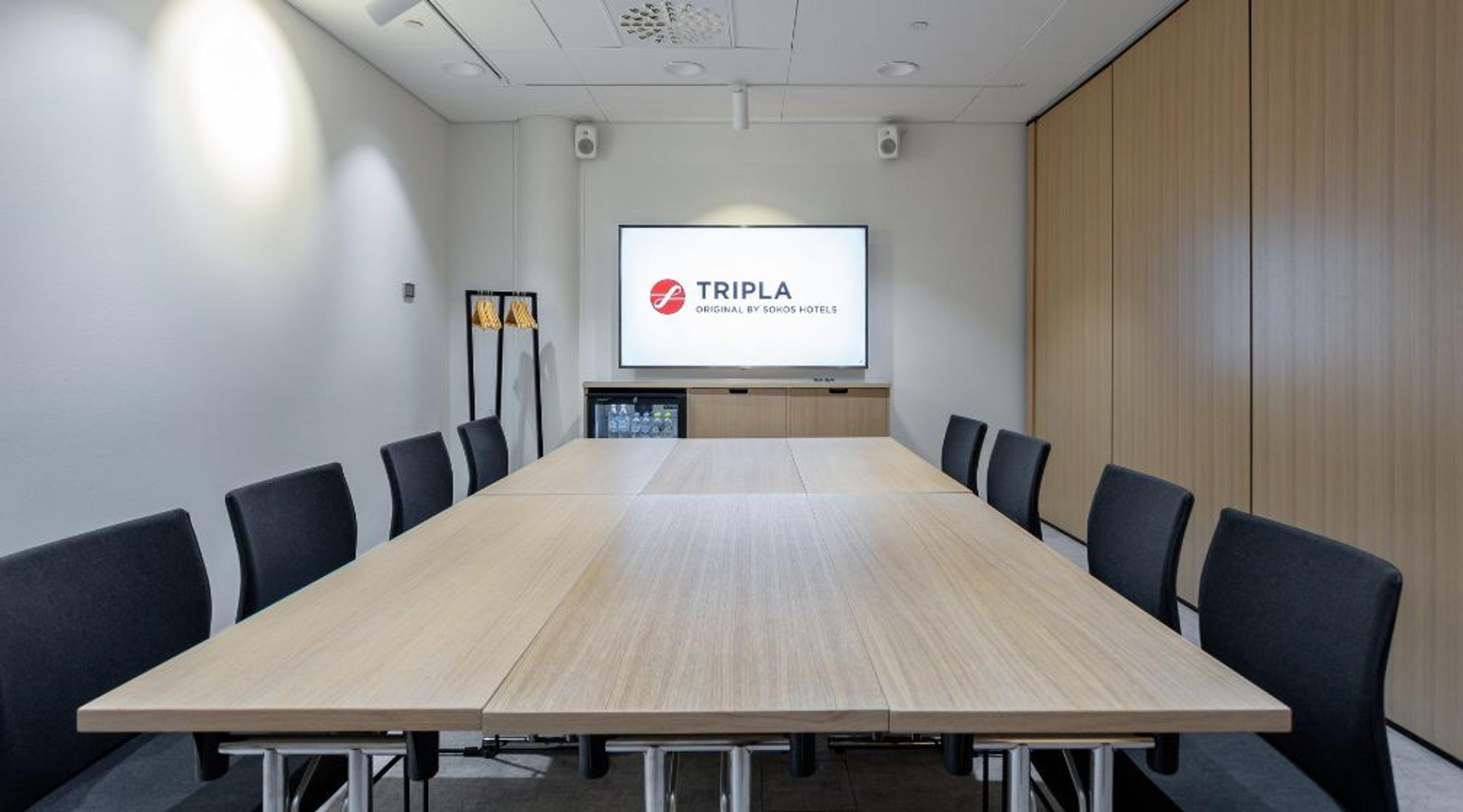 Modernt mötesrum med ljust träbord och svarta stolar. Framme finns en stor skärm, på sidan träpaneler.