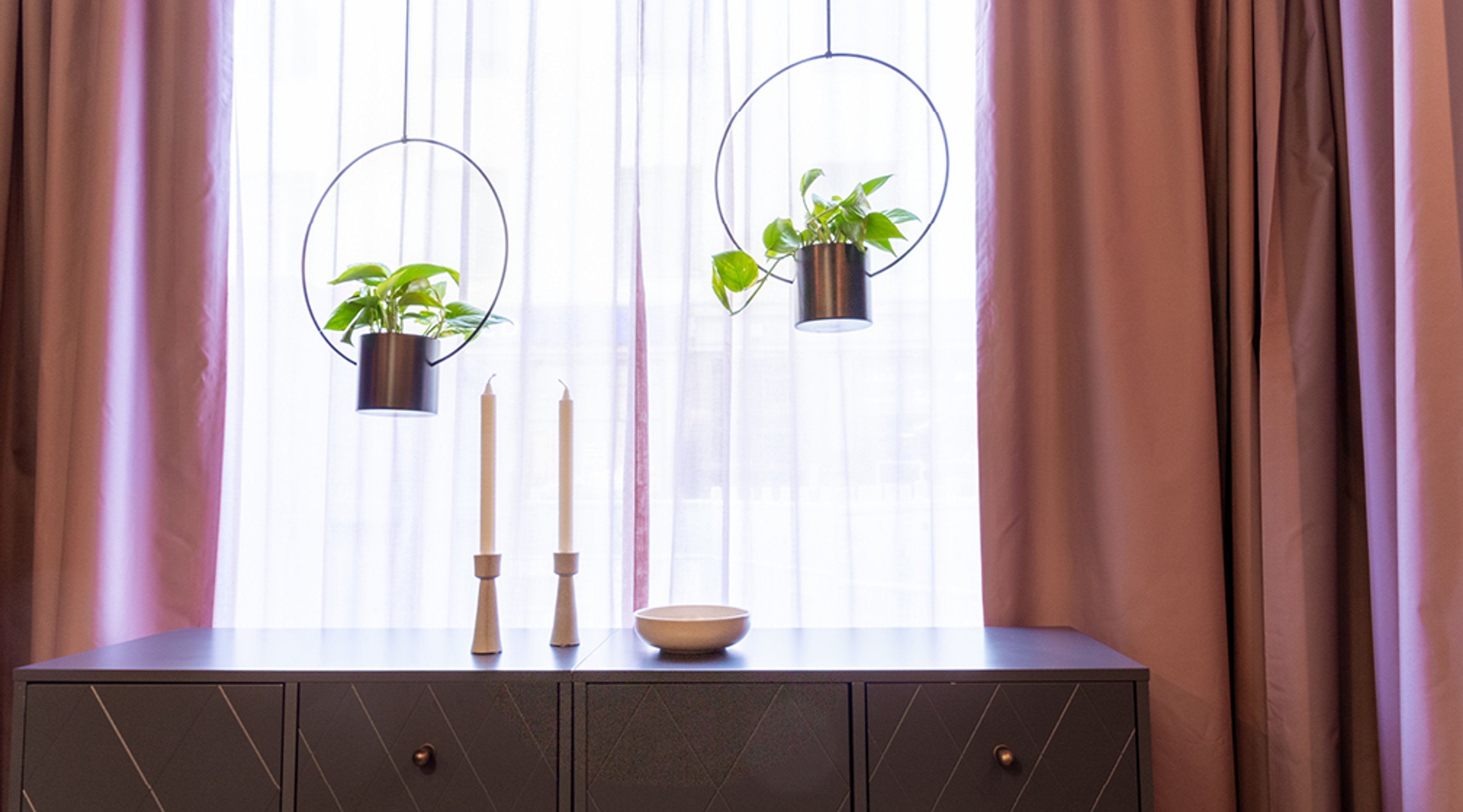 Lipasto ikkunan edessä, jossa kaksi valkoista kynttilää, puinen kulho ja kaksi riippuvaa viherkasvia metallisissa ruukuissa.