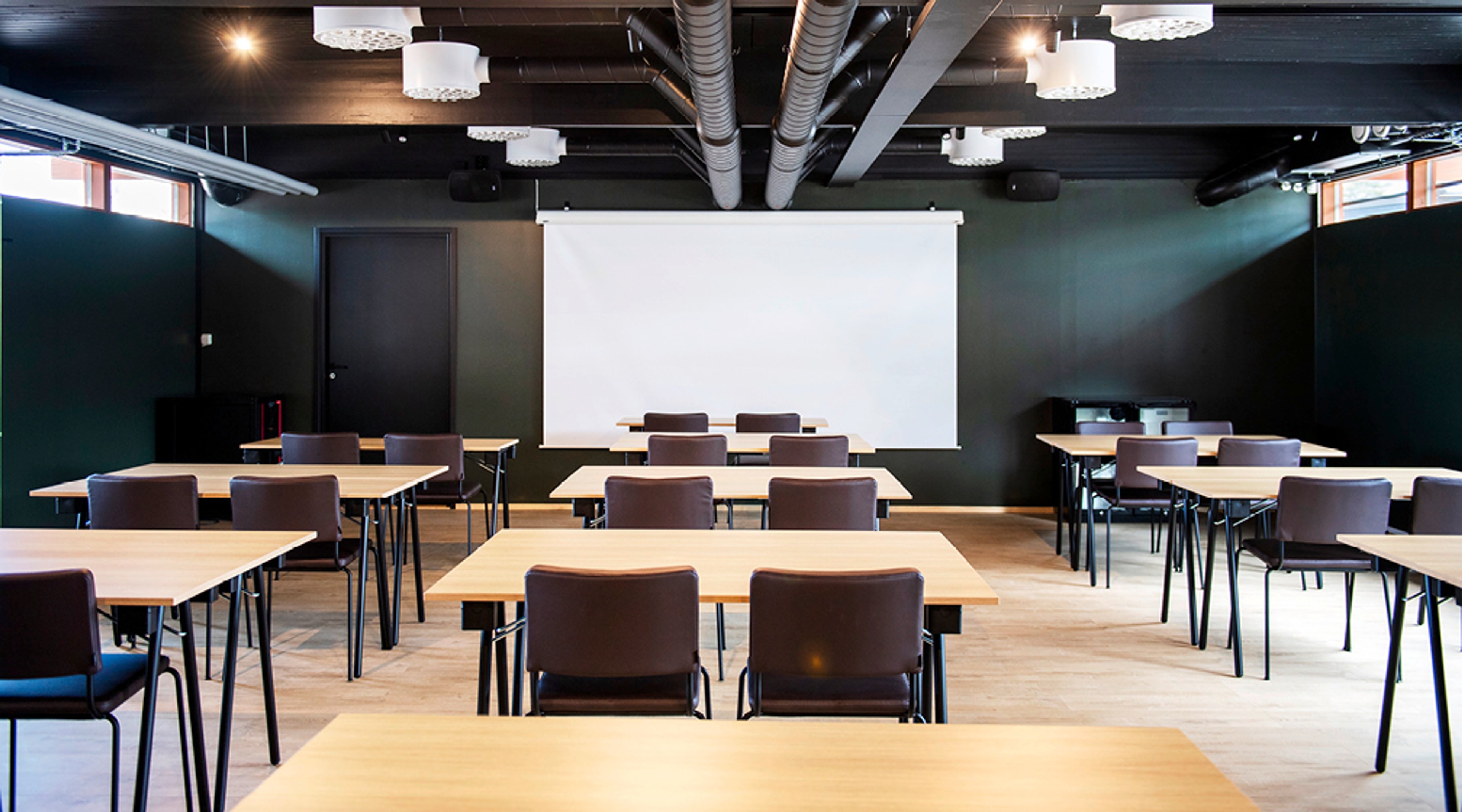 Moderni kokoustila mustilla seinillä ja puisella lattialla, jossa on vaaleita pöytiä ja ruskeita tuoleja. Valkoinen projektiokangas edessä.