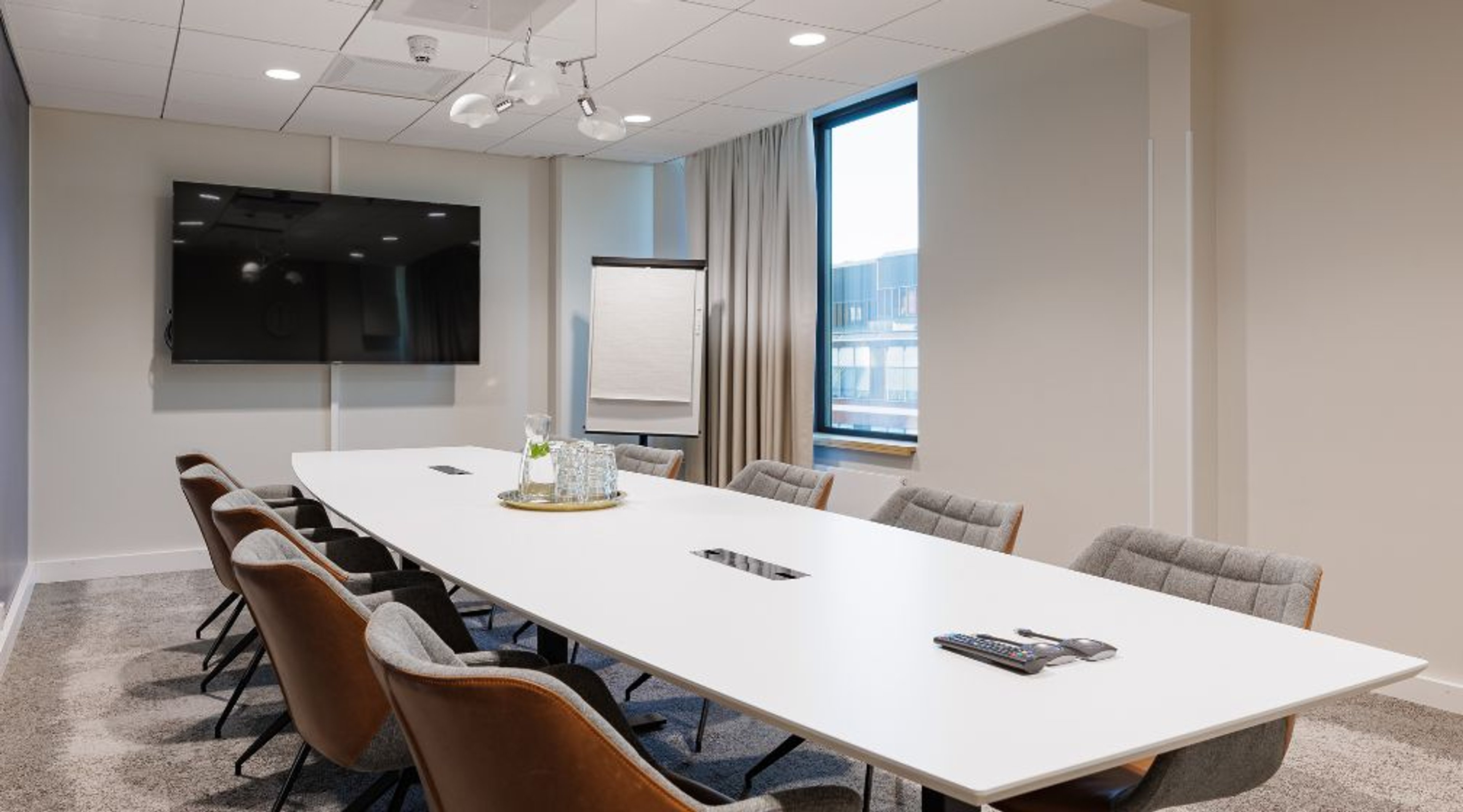 Moderni neuvotteluhuone, jossa pitkä valkoinen pöytä, ruskeita ja harmaita tuoleja, seinälle asennettu televisio ja fläppitaulu. Huoneessa on ikkuna ja tasainen kokolattiamatto.
