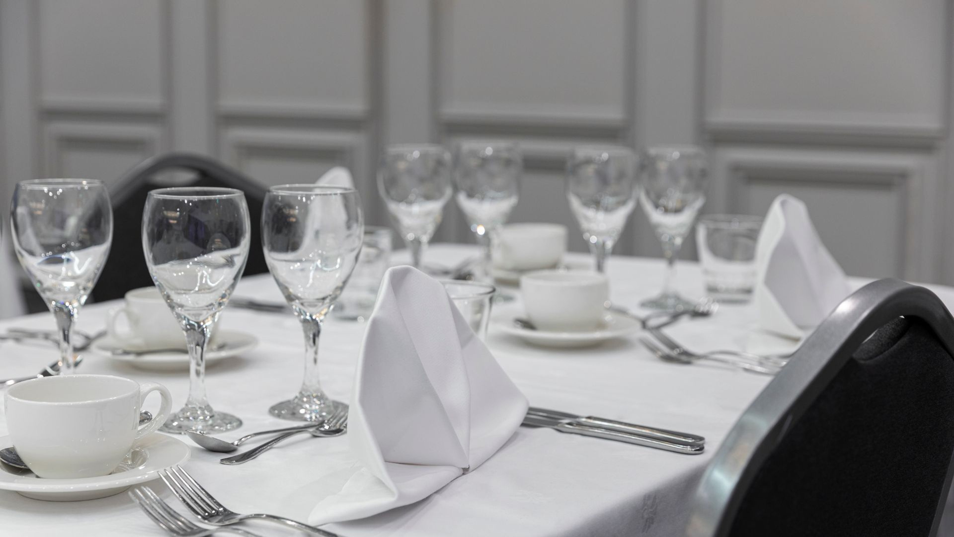 Elegantisti katettu pöytä valkoisella pöytäliinalla, viinilaseja, taitettu lautasliina, kahvikuppi ja ruokailuvälineet.