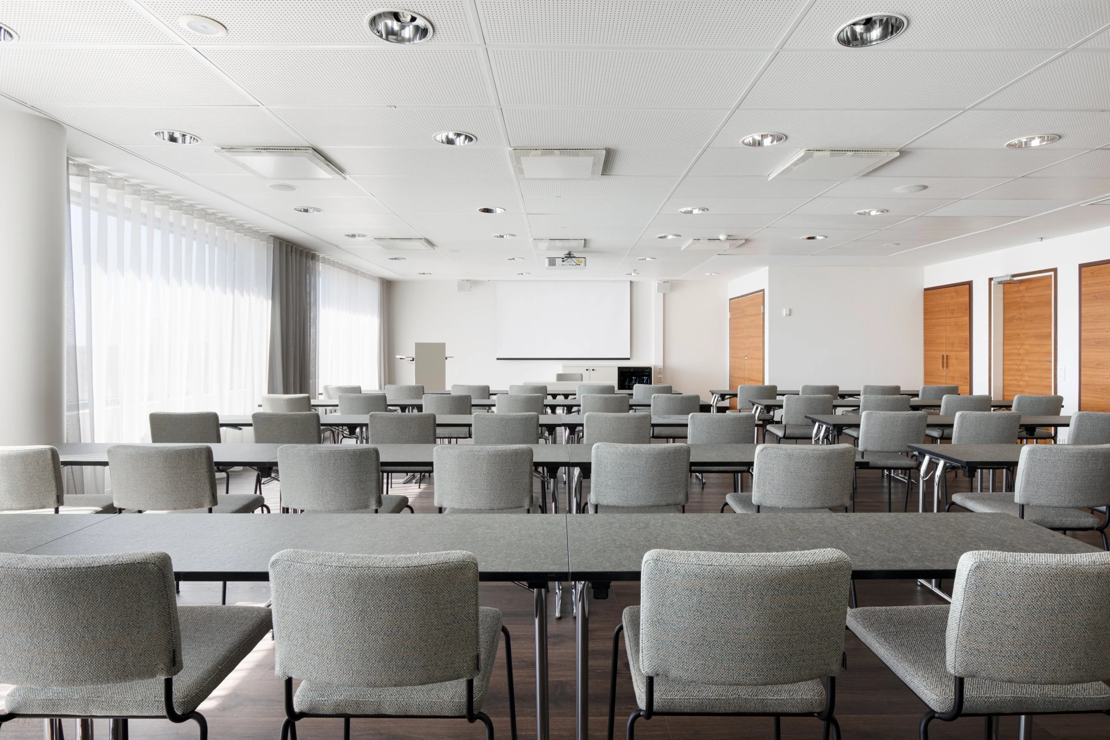 Moderni kokoushuone harmailla tuoleilla, valkoisilla seinillä ja puisilla ovilla. Edessä valkoinen projektorikangas ja isot ikkunat vasemmalla.