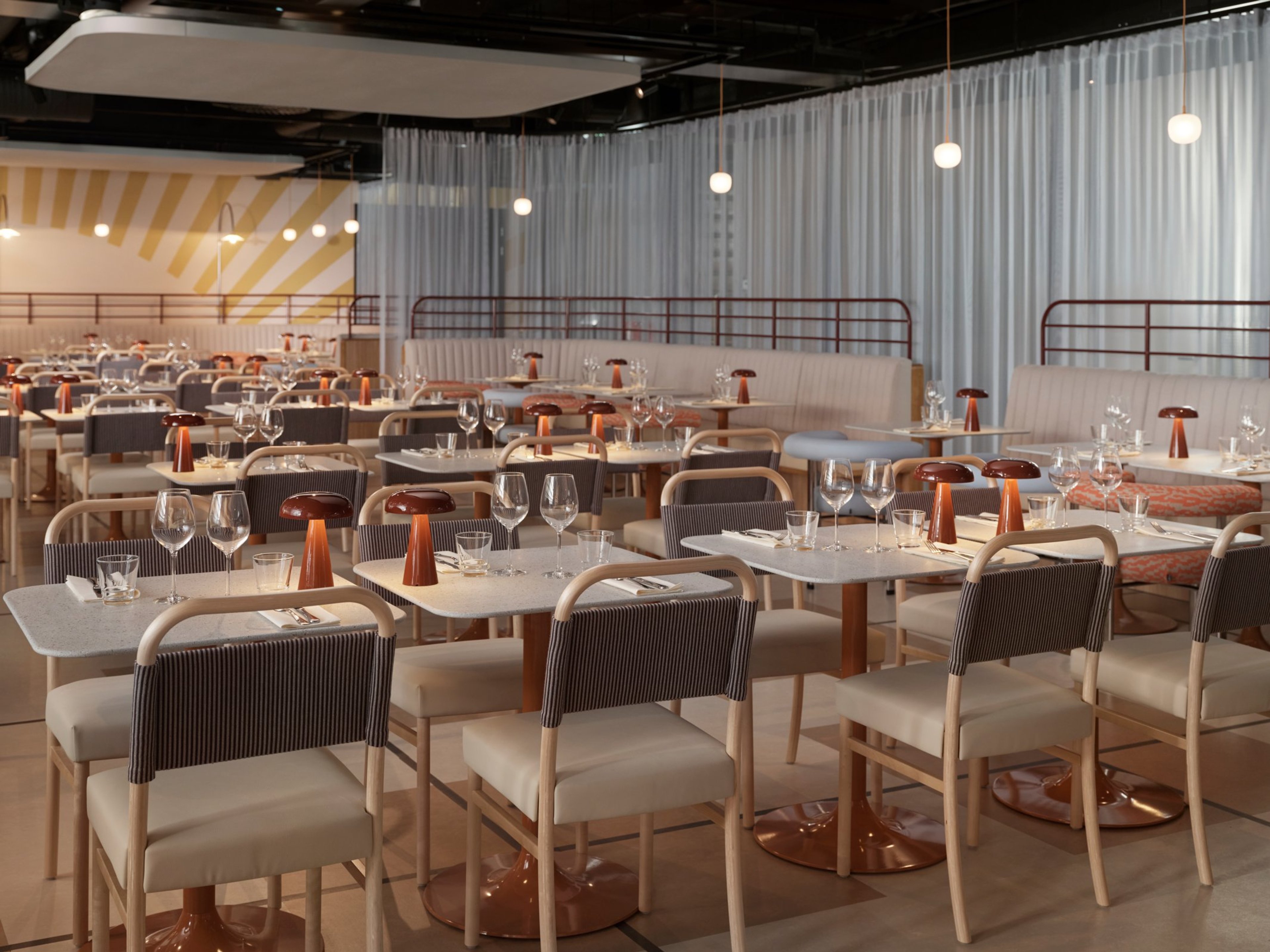 Moderni ravintola vaalein pöydin ja tuolein. Pöydillä on laseja ja kuparinväriset pöytävalaisimet. Taustalla valkoisia verhoja ja seinäpenkkejä.