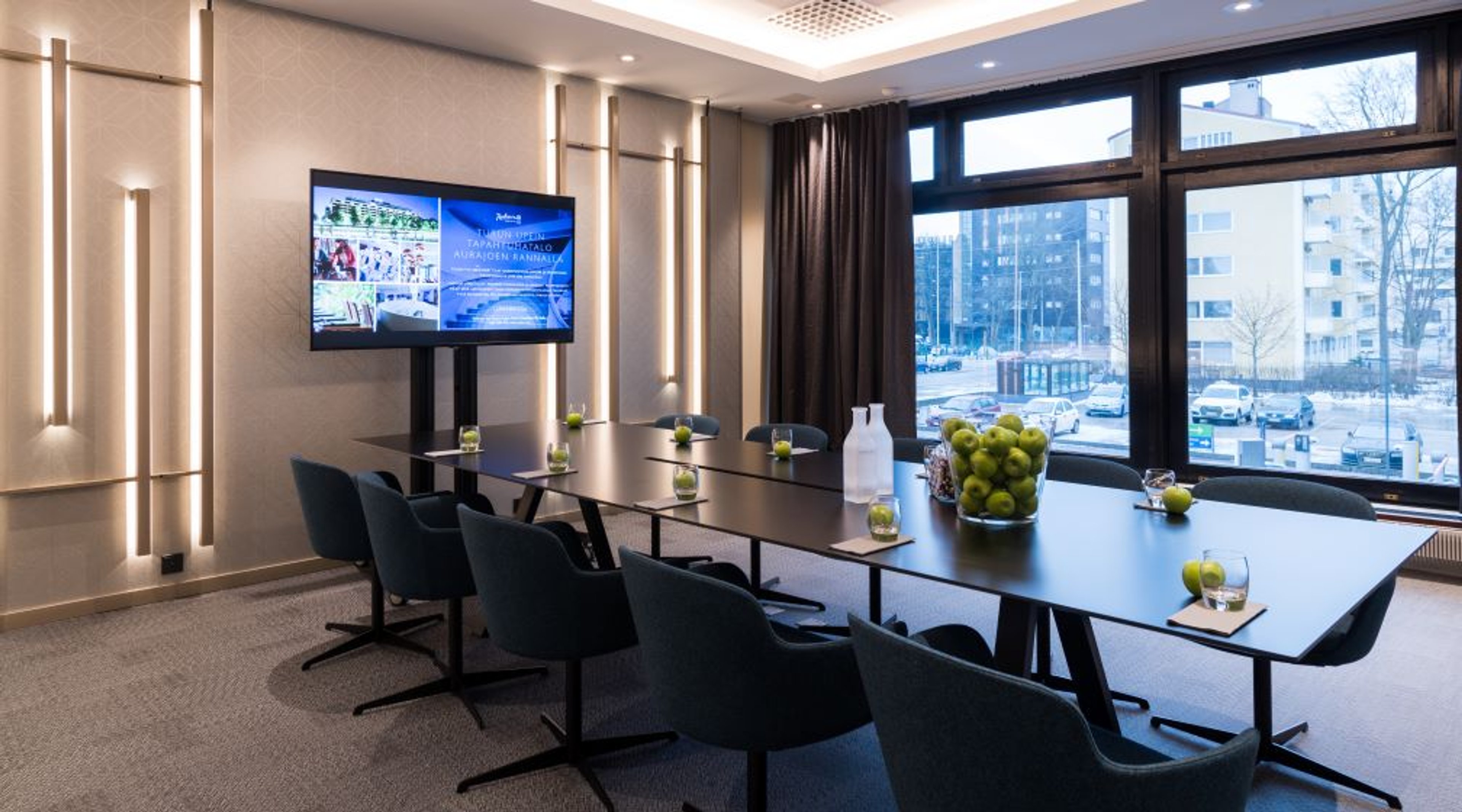 Modern conference room with black table, wall-mounted screen, green apples on the table and large windows overlooking city view.