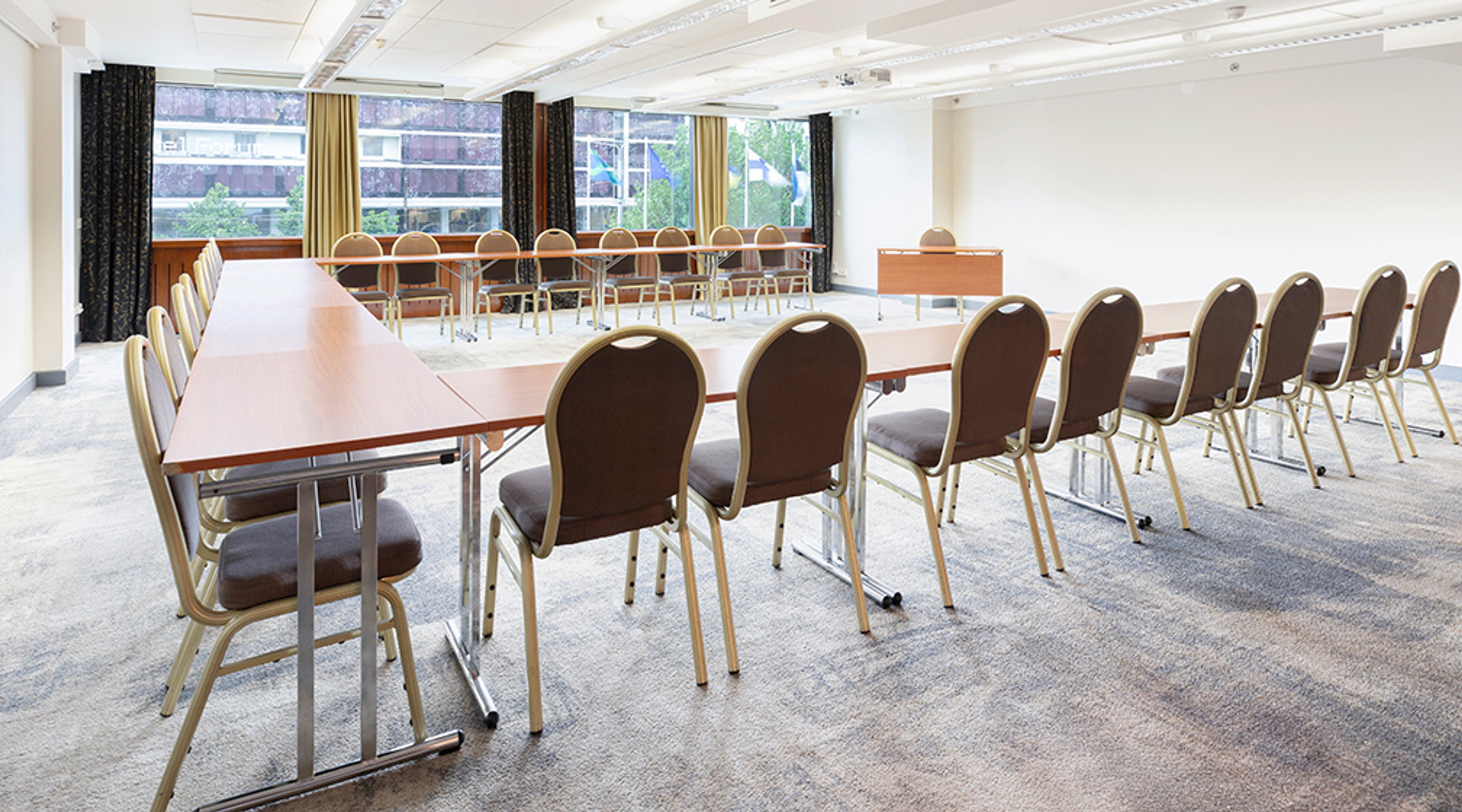 Conference room with long tables and chairs arranged in U-shape, large windows in background and curtained area. Floor is level with low-pile carpet.