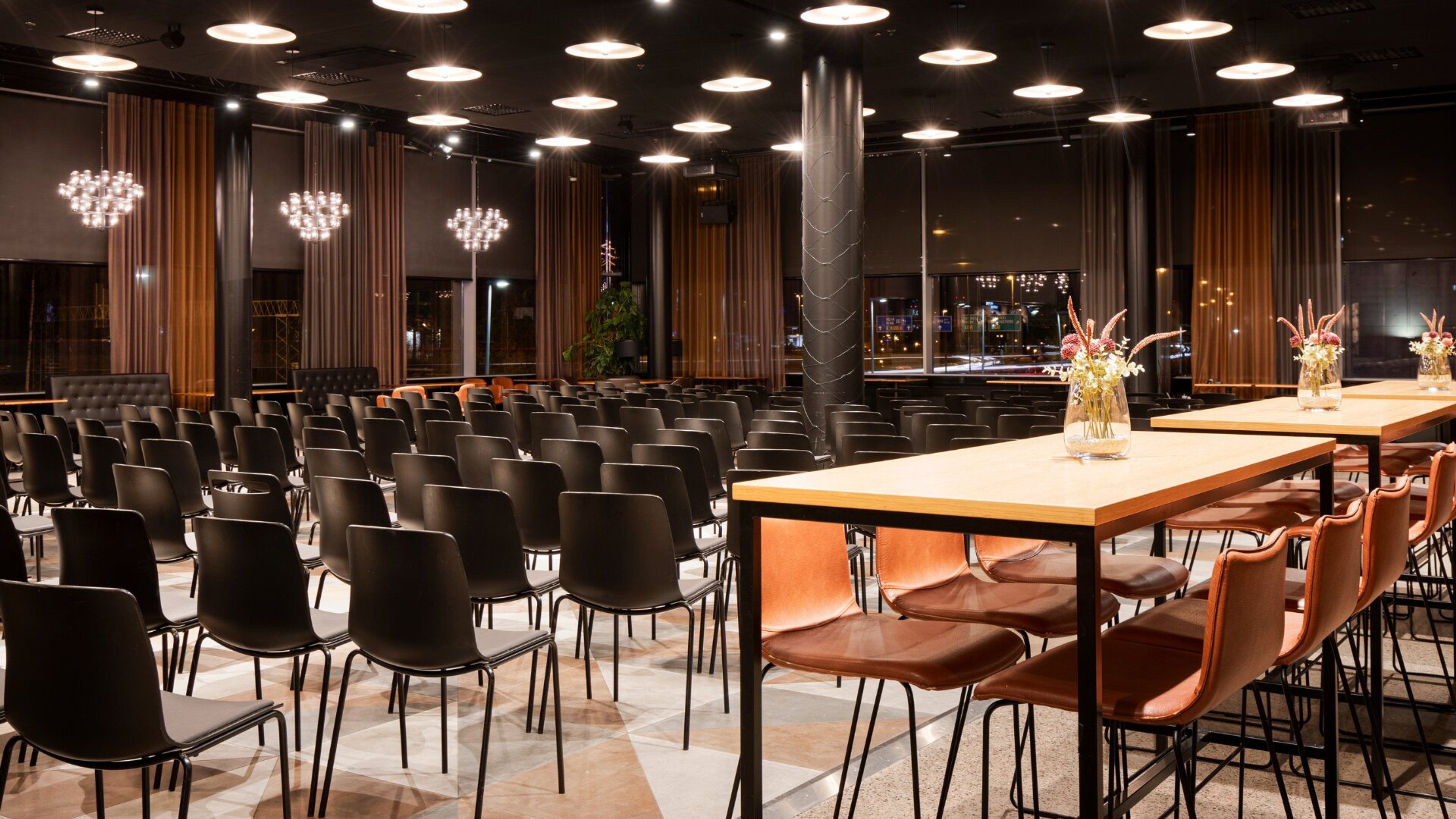 Moderni tapahtumatila riveittäin järjestetyillä mustilla tuoleilla ja edessä paneelipöydillä. Tilassa isot ikkunat, lämpimät verhot ja tasainen laattalattia.