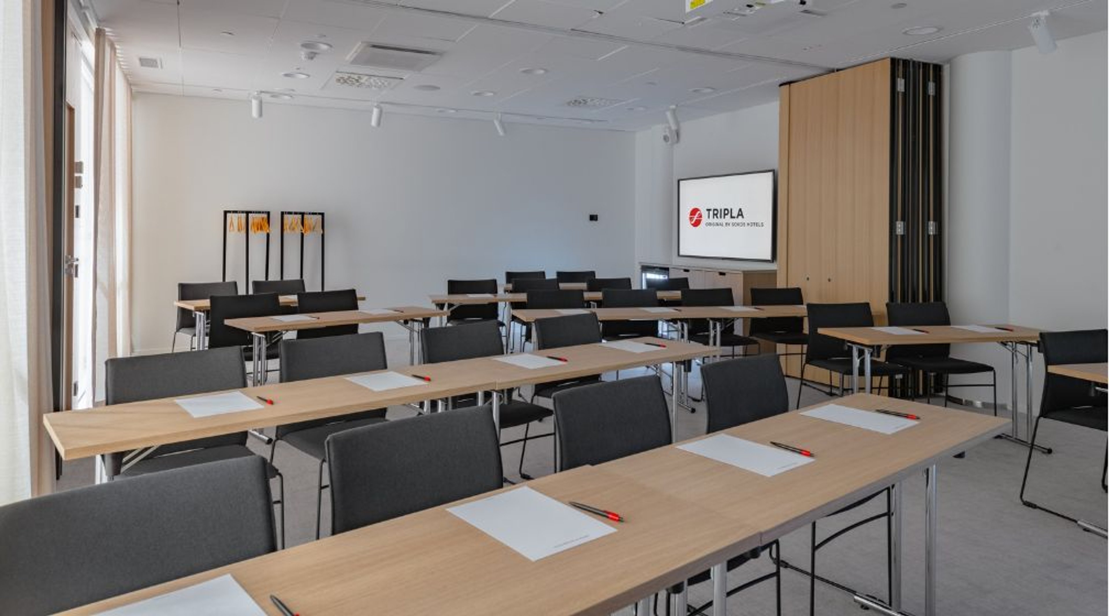 Modern meeting room with rows of tables and chairs. Projection screen visible at front and light-colored walls. Floor has hard surface and room features good lighting.