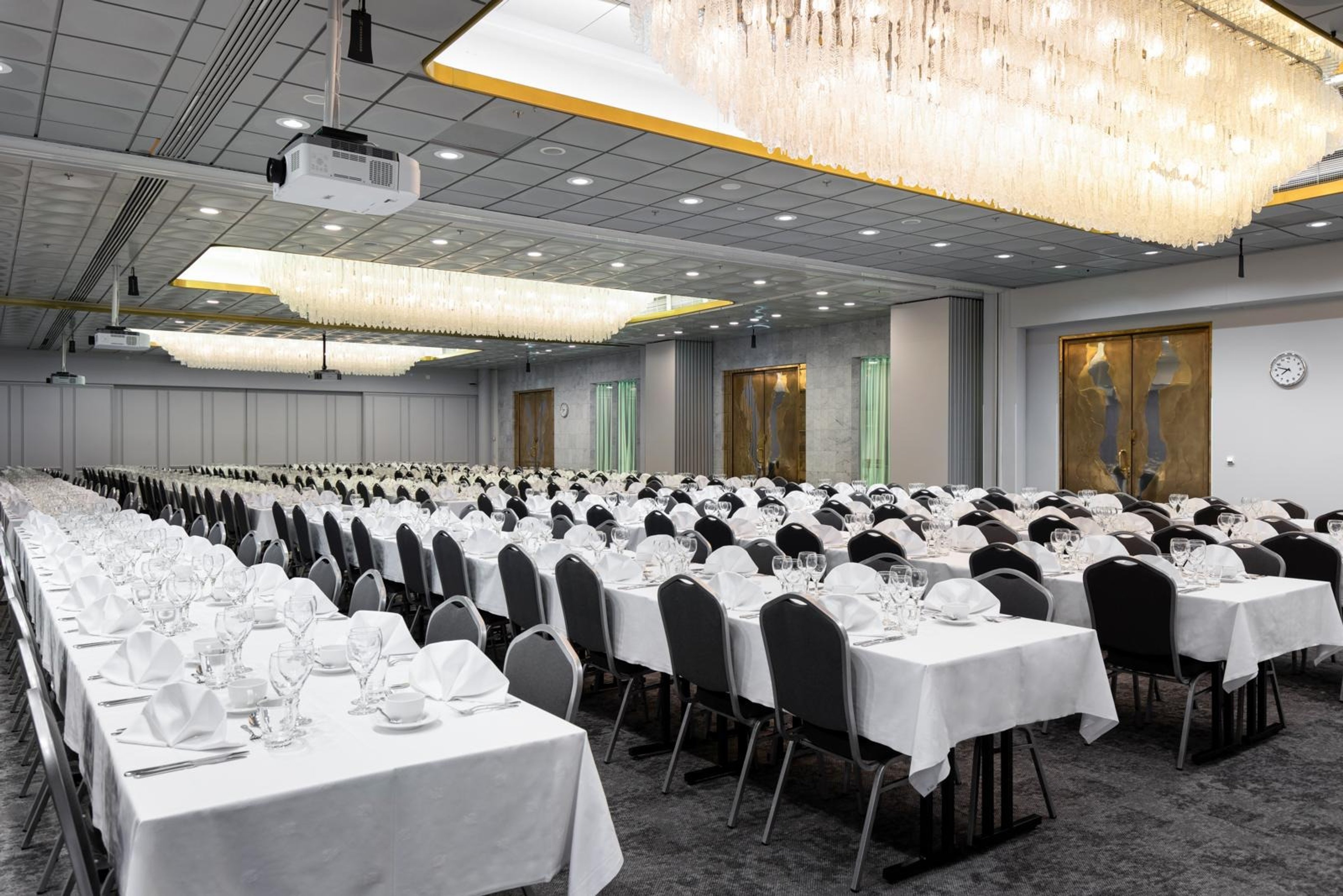 Juhlasali, jossa pitkät pöydät valkoisilla liinoilla ja mustilla tuoleilla. Kattokruunut ja projektorit katossa, tasainen lattia.