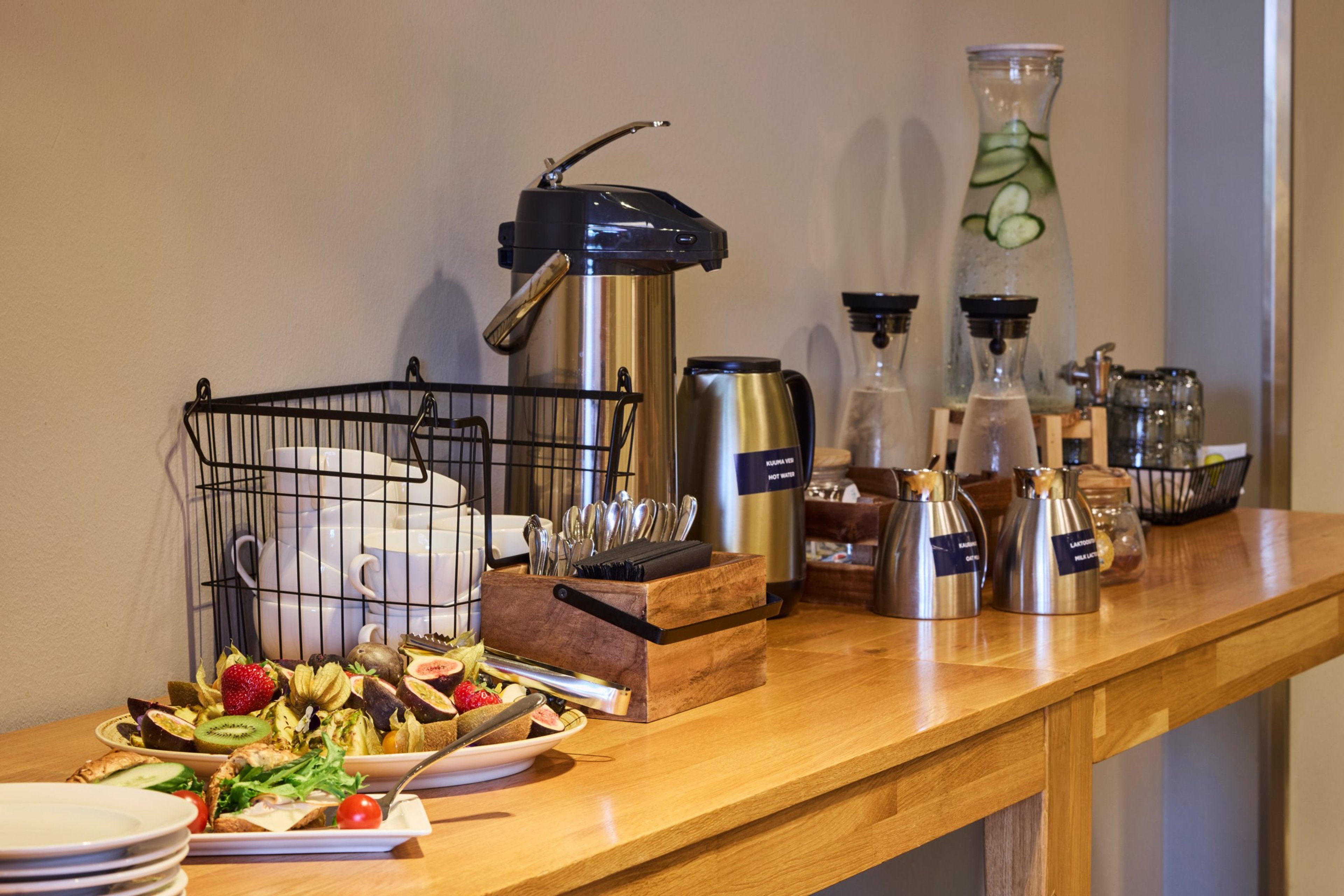 Kahvi- ja juomapiste puisella pöydällä. Termospannu, mukeja korissa, hedelmiä lautasella, vesikannu kurkun viipaleilla ja muita tarjoiluastioita.