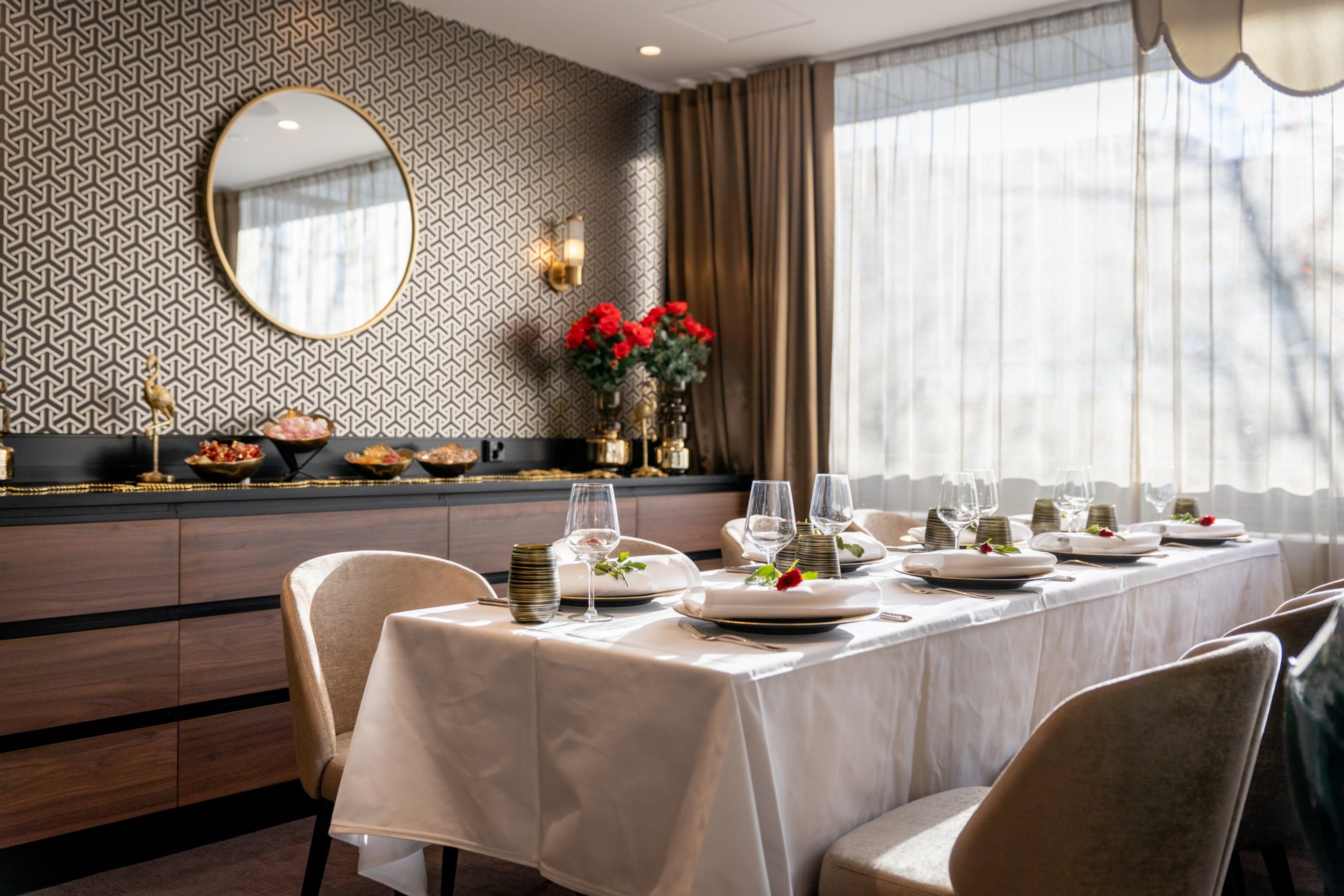 Elegantti ruokailutila, jossa katettu pöytä valkoisella liinalla, taustalla kuviollinen tapetti ja pyöreä peili. Sivupöydällä on tarjoiluja ja punaisia kukkia.