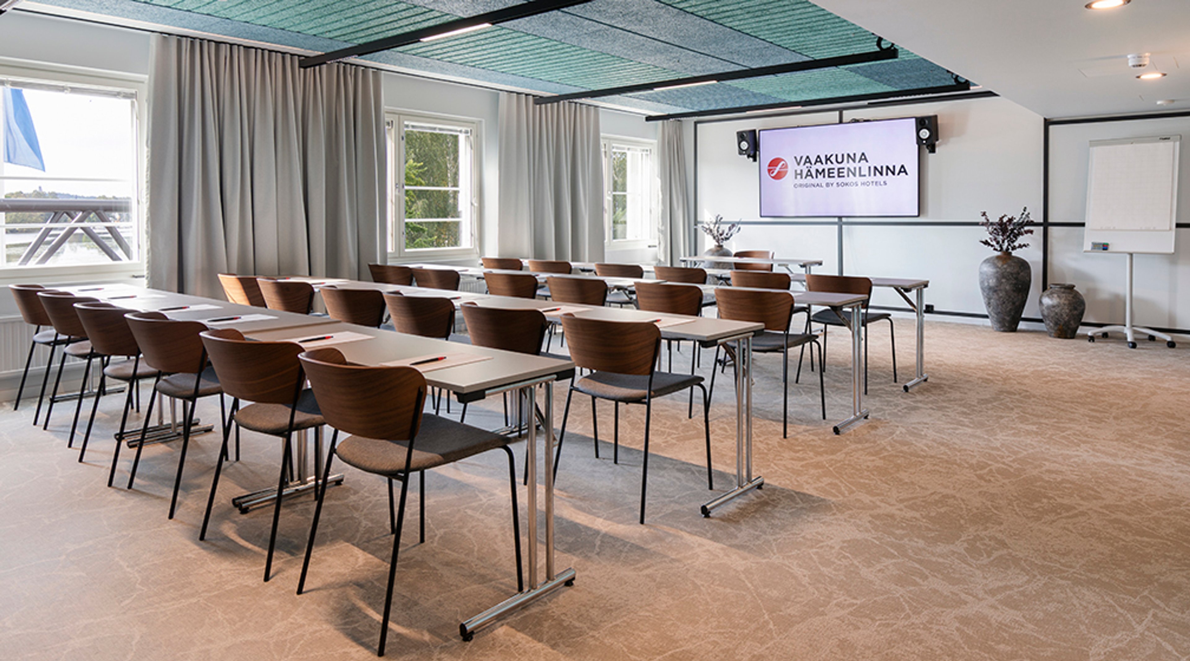 Valoisa kokoustila, jossa on ruskeat tuolit ja pöydät luokkamuodostelmassa. Tilassa on ikkunat, näyttö edessä ja tasainen lattia.