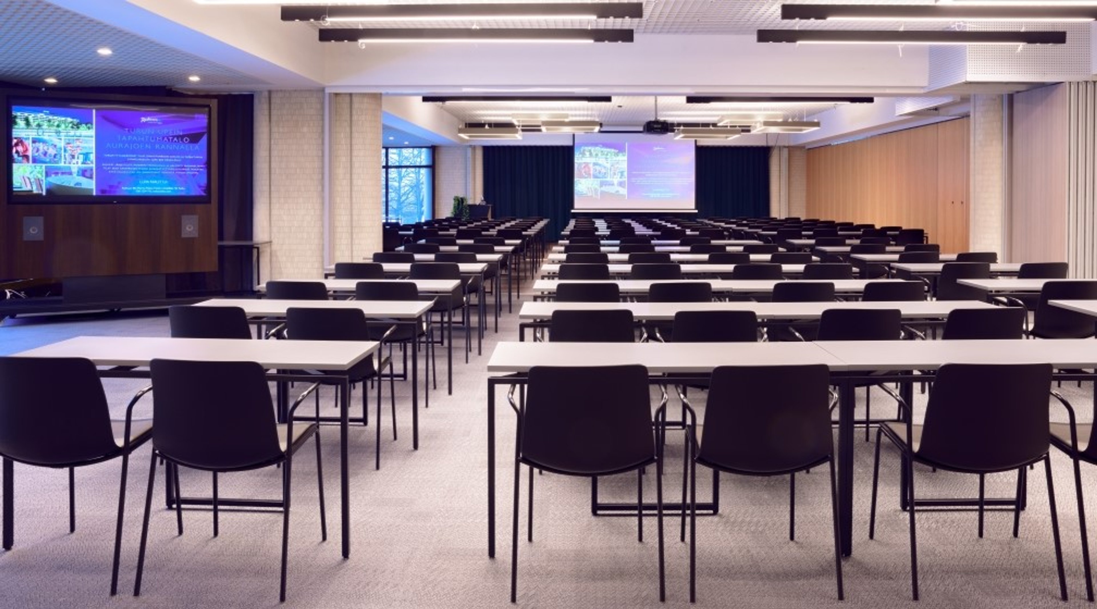 Suuri luentosali järjestettynä luokkahuonetyyliin valkoisilla pöydillä ja mustilla tuoleilla. Edessä kaksi esitysruutua ja tasainen lattiapinta.