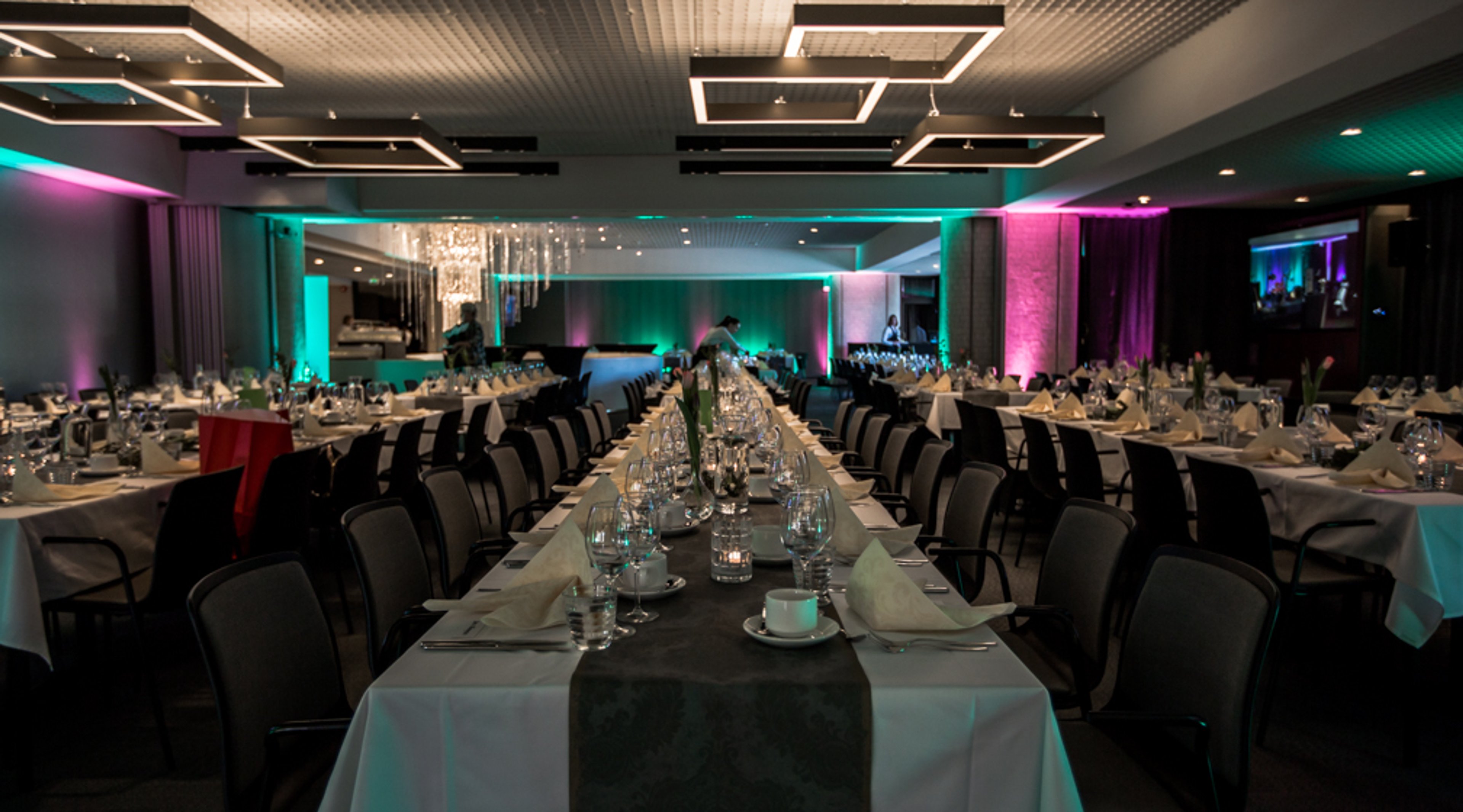 Juhlasali koristeellisella valaistuksella, pitkät pöydät valmiina ruokailua varten. Turkoosit ja pinkit valot seinillä.