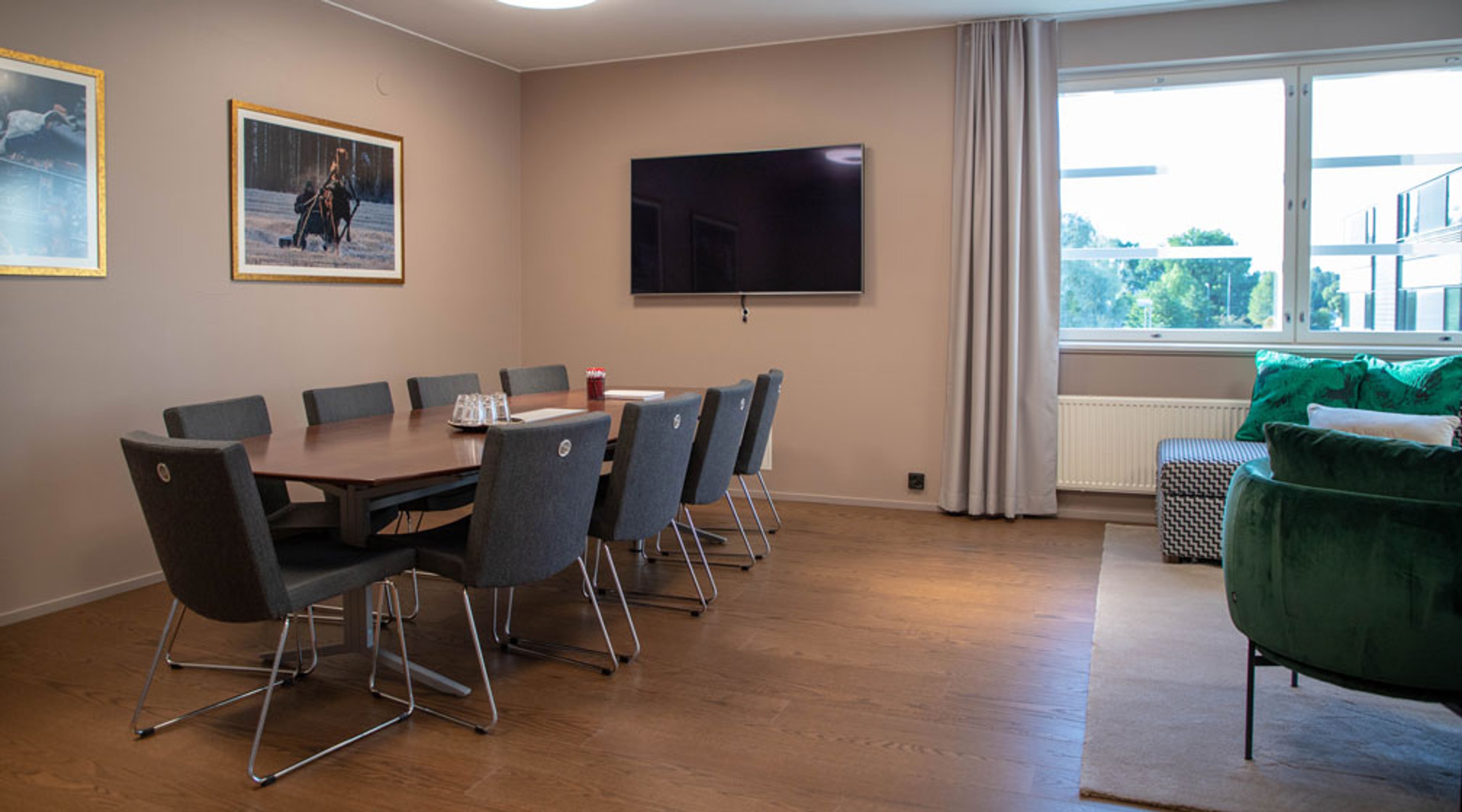 Kokoushuone, jossa puinen neuvottelupöytä harmaine tuoleineen, seinällä tv ja tauluja, isot ikkunat ja vihreä sohva nurkassa. Puulattia.