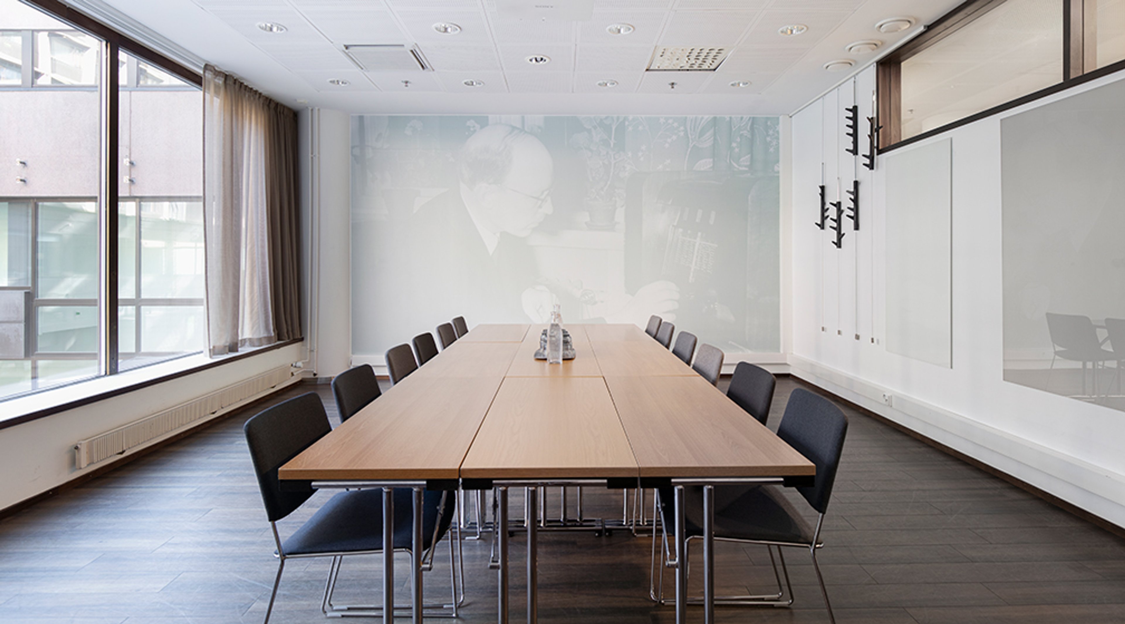 Moderni neuvotteluhuone, jossa pitkä puinen pöytä, mustia tuoleja, suuri ikkunaseinä ja historiallinen valokuva seinällä.
