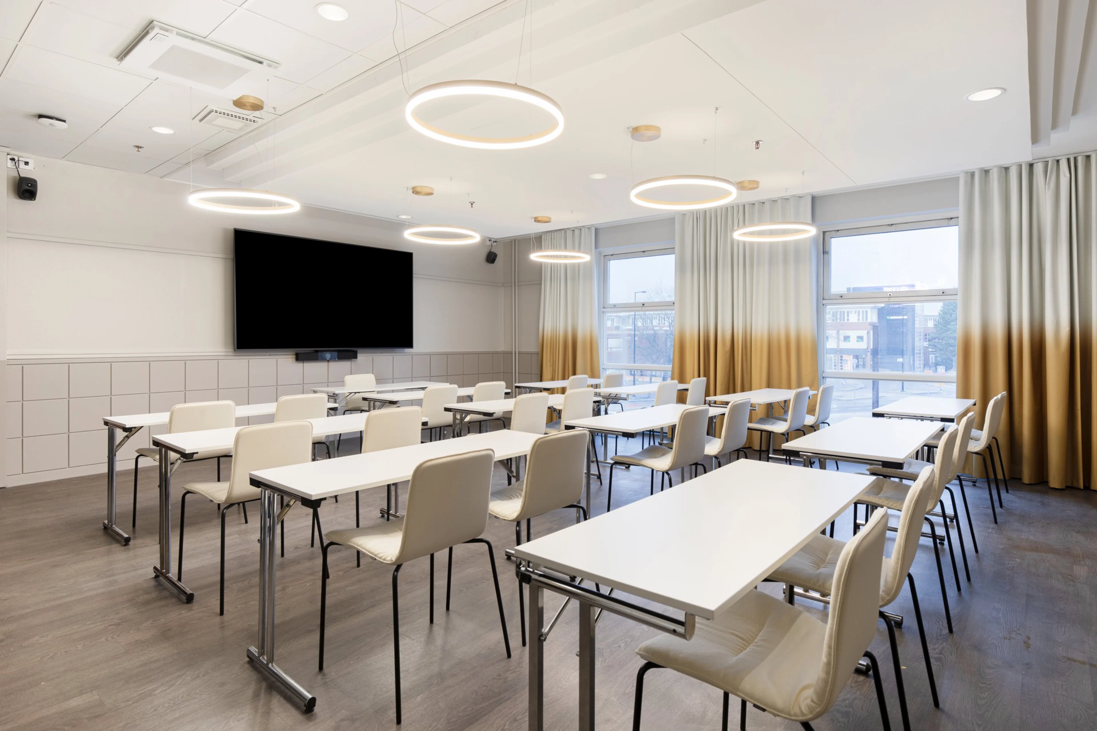 Moderni luokkahuone valkoisilla pöydillä ja tuoleilla, suurella näytöllä edessä, rengasmaisilla valaisimilla ja ombré-verhoilla.