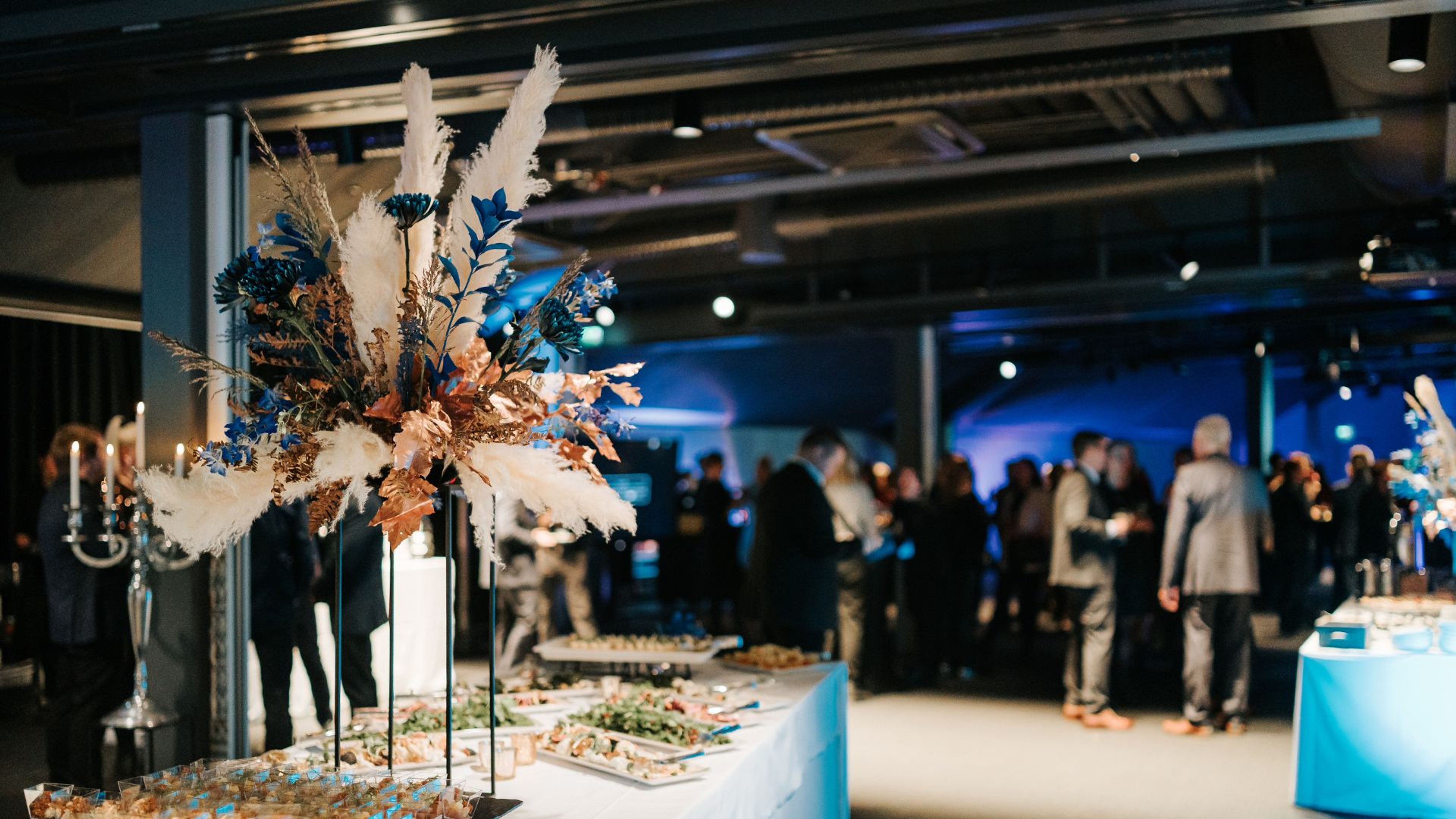 Juhlatila sinisessä valaistuksessa, jossa noutopöytä koristeellisilla kukka-asetelmilla. Taustalla vieraita tasaisella lattialla.