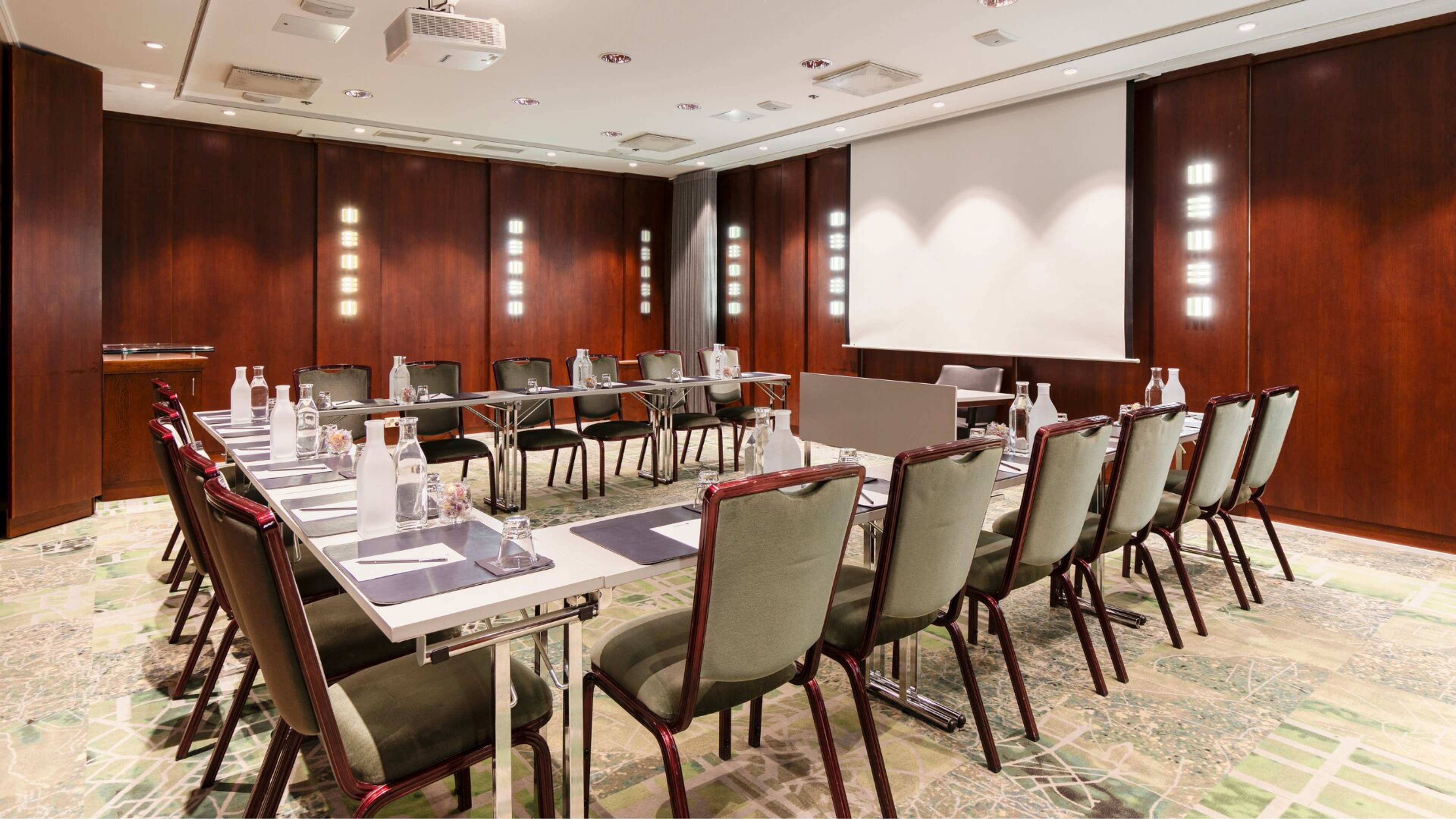 Kokoushuone, jossa mahonkipaneelit seinillä, U-muotoinen pöytäjärjestely, vihreät tuolit ja valkokangas. Tasainen lattia, kirkas valaistus.