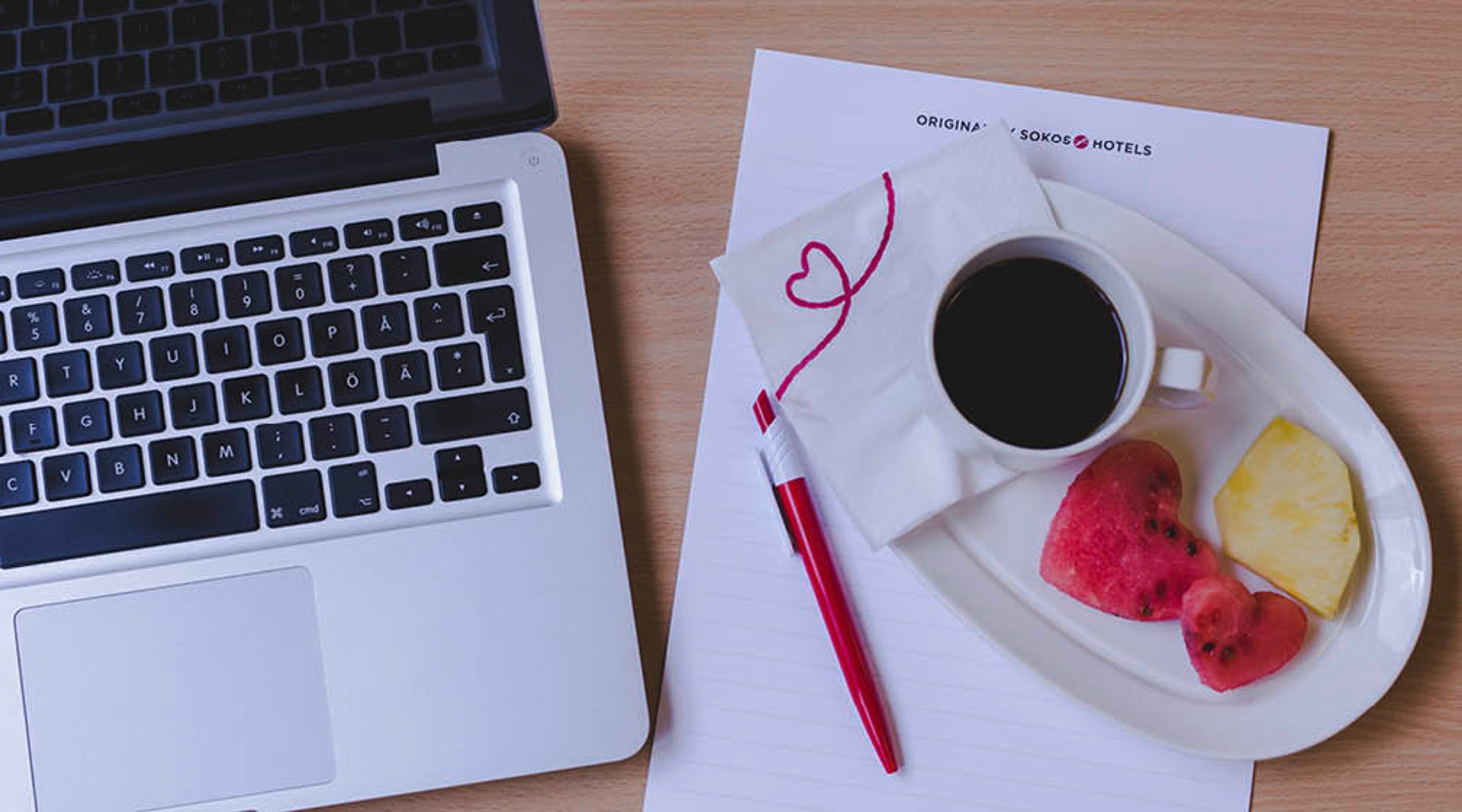 Kannettava tietokone ja valkoisella lautasella kahvikuppi, vesimelonia ja ananasta puisella pöydällä. Punainen kynä paperilla.