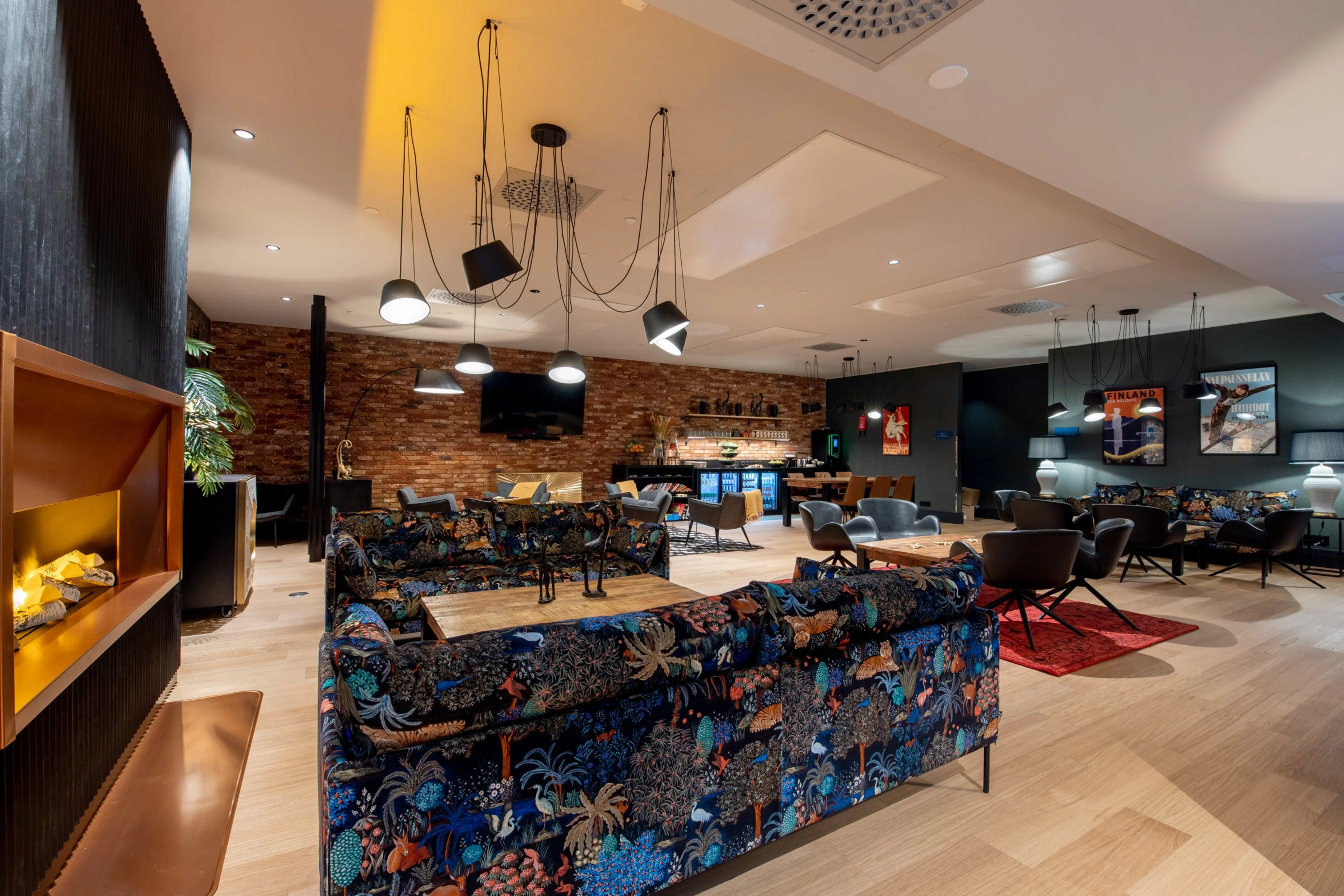 Moderni oleskelutila, jossa on värikkäitä sohvia, puiset pöydät, tiiliseinä, laskeva valaisinjärjestelmä ja kovapuinen lattia.