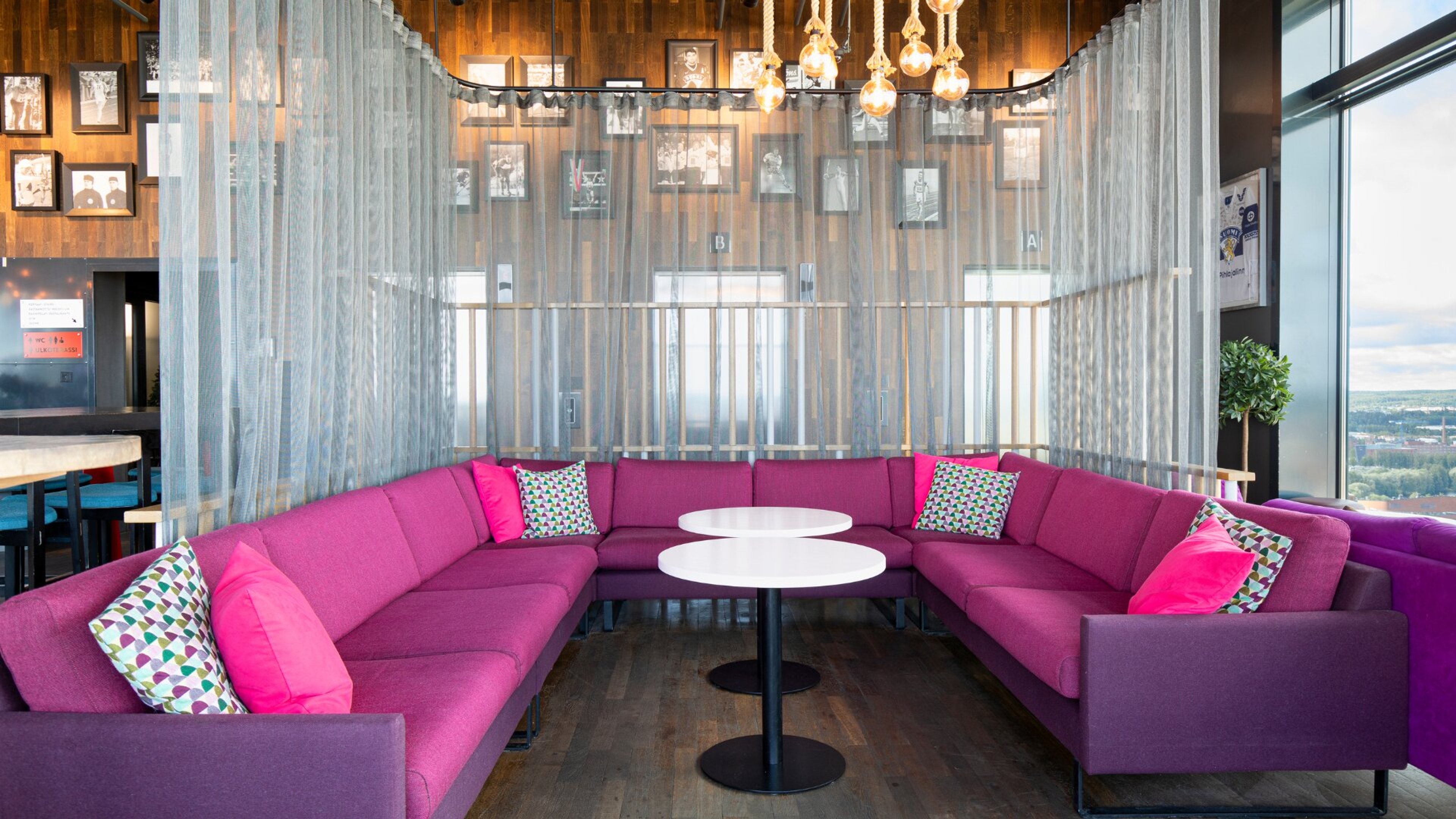 Loungealue, jossa pinkki kulmasohva, kaksi pyöreää pöytää, puinen lattia, läpikuultavat verhot ja roikkuvat lamput. Taustalla valokuvia puupaneeliseinällä.