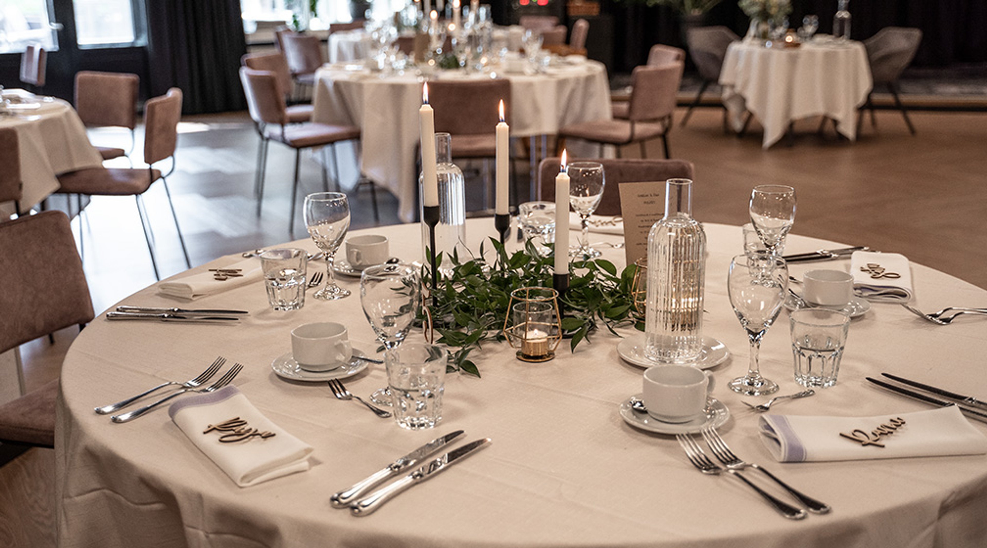 Juhlallisesti katettu pyöreä pöytä kynttilöillä, vihreillä koristeilla ja lautasliinoilla. Taustalla ravintolasali, jossa lisää pöytiä.