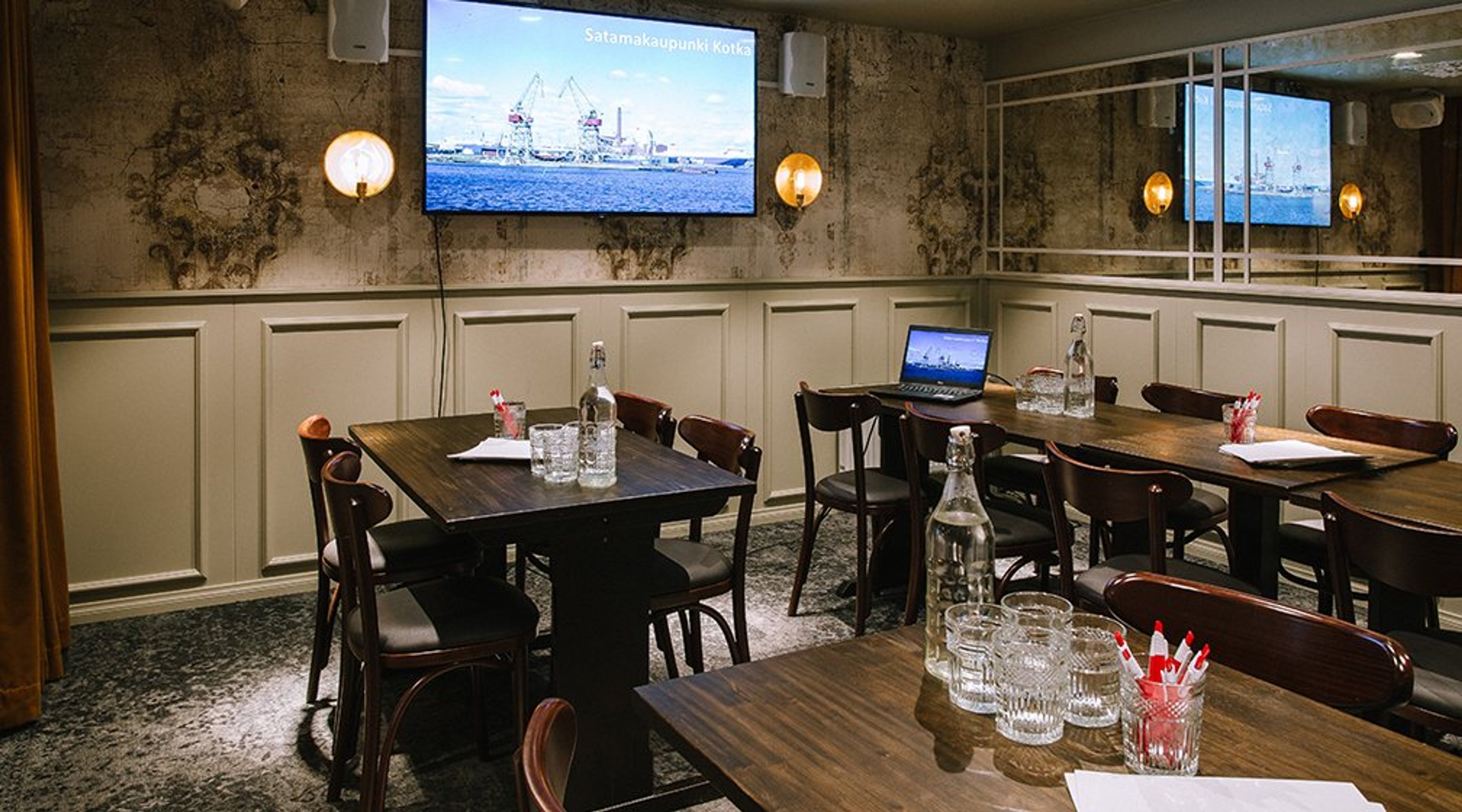 Ravintolan kokoustila, jossa tummat pöydät, tuolit ja seinällä iso näyttö. Tyylikkäät seinävalaisimet ja panelointi seinissä.