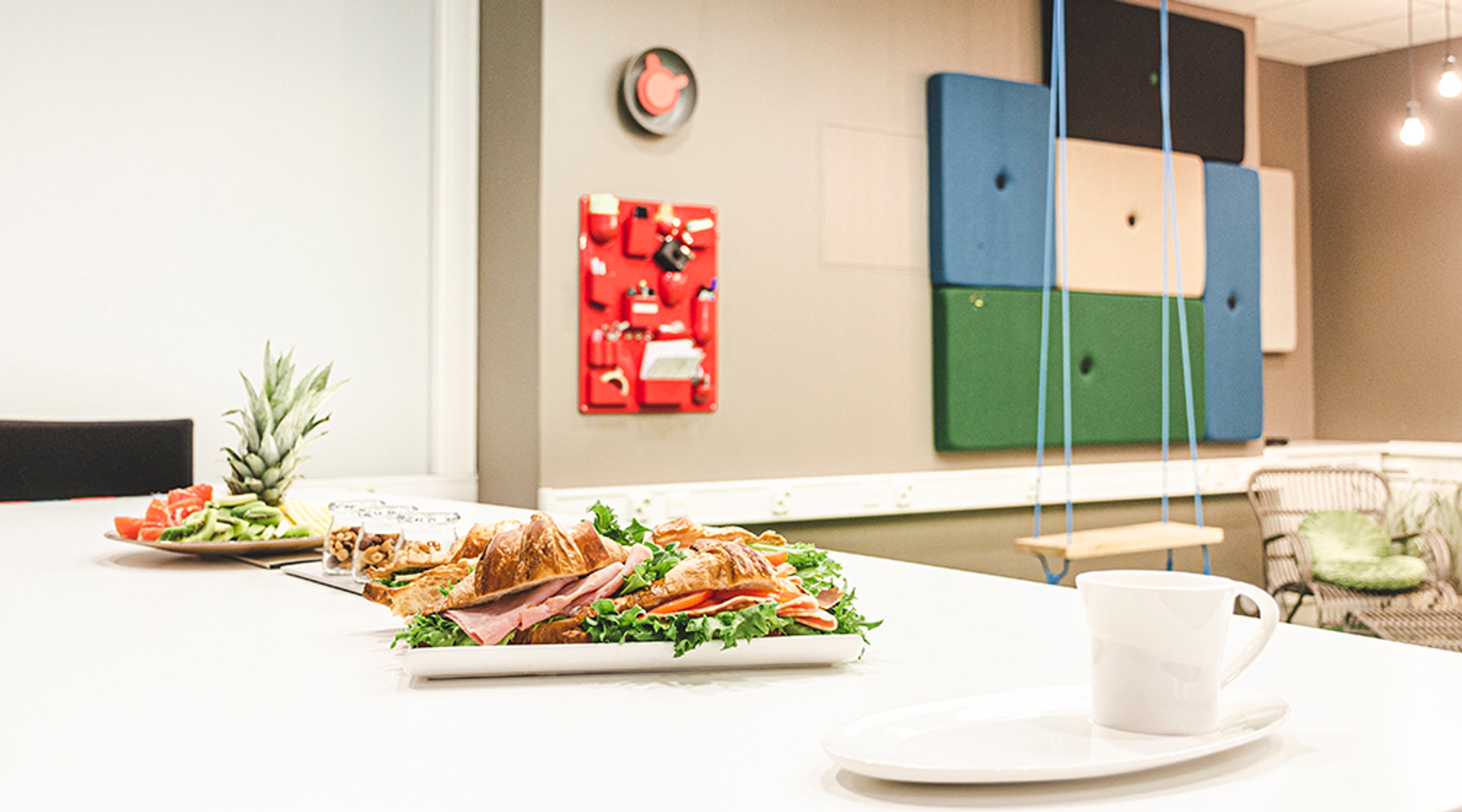 Valkoinen pöytä, jossa tarjoiltuna croissant-voileipä, hedelmälautanen ja kahvikuppi. Taustalla värikäs akustiikkapaneeliseinä ja kello.