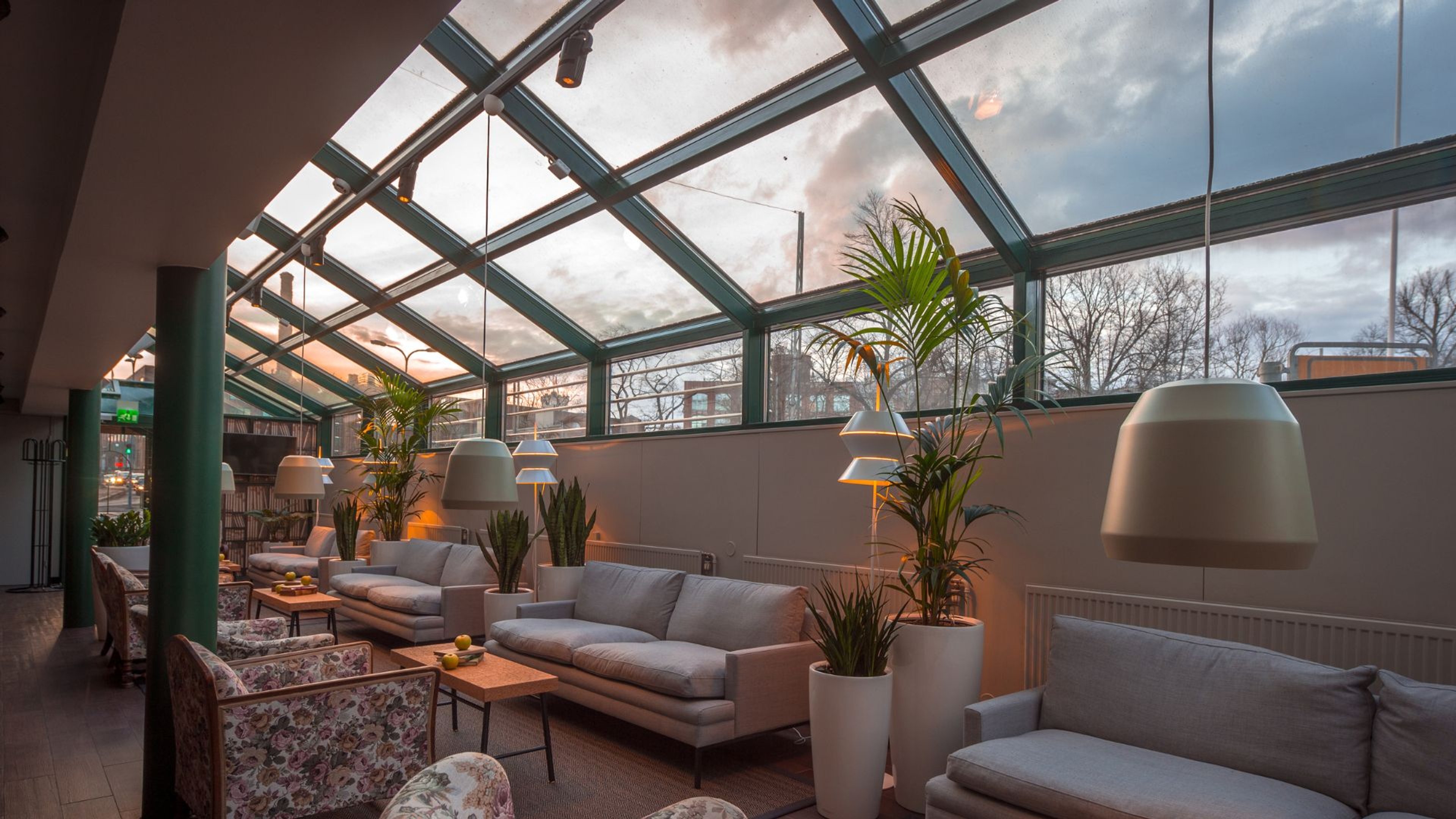 Viihtyisä lasikattoisessa oleskelutila, jossa harmaita sohvia, kukkakuvioisia nojatuoleja ja viherkasveja. Luonnonvaloa tulvii sisään ja taustalla näkyy puita.