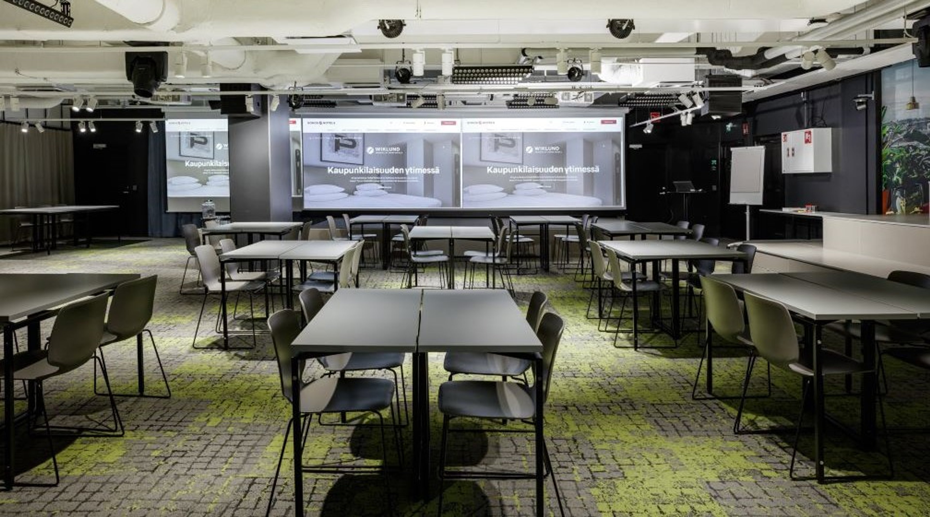 Moderni kokoustila, jossa useita pöytiä ja tuoleja. Kolme näyttöä etuseinällä ja tasainen, kuviollinen lattia vihreällä ja harmaalla sävyllä.