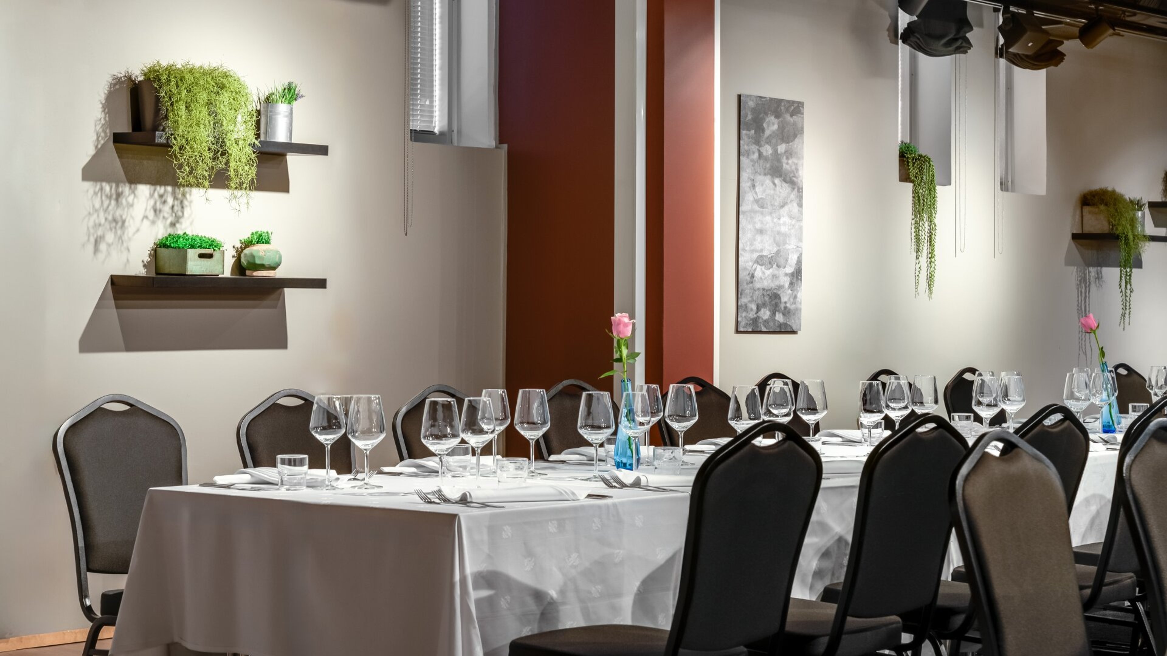 Bright dining area with white tablecloth-covered table and dark chairs. Wall shelves display greenery and table is set with glassware.