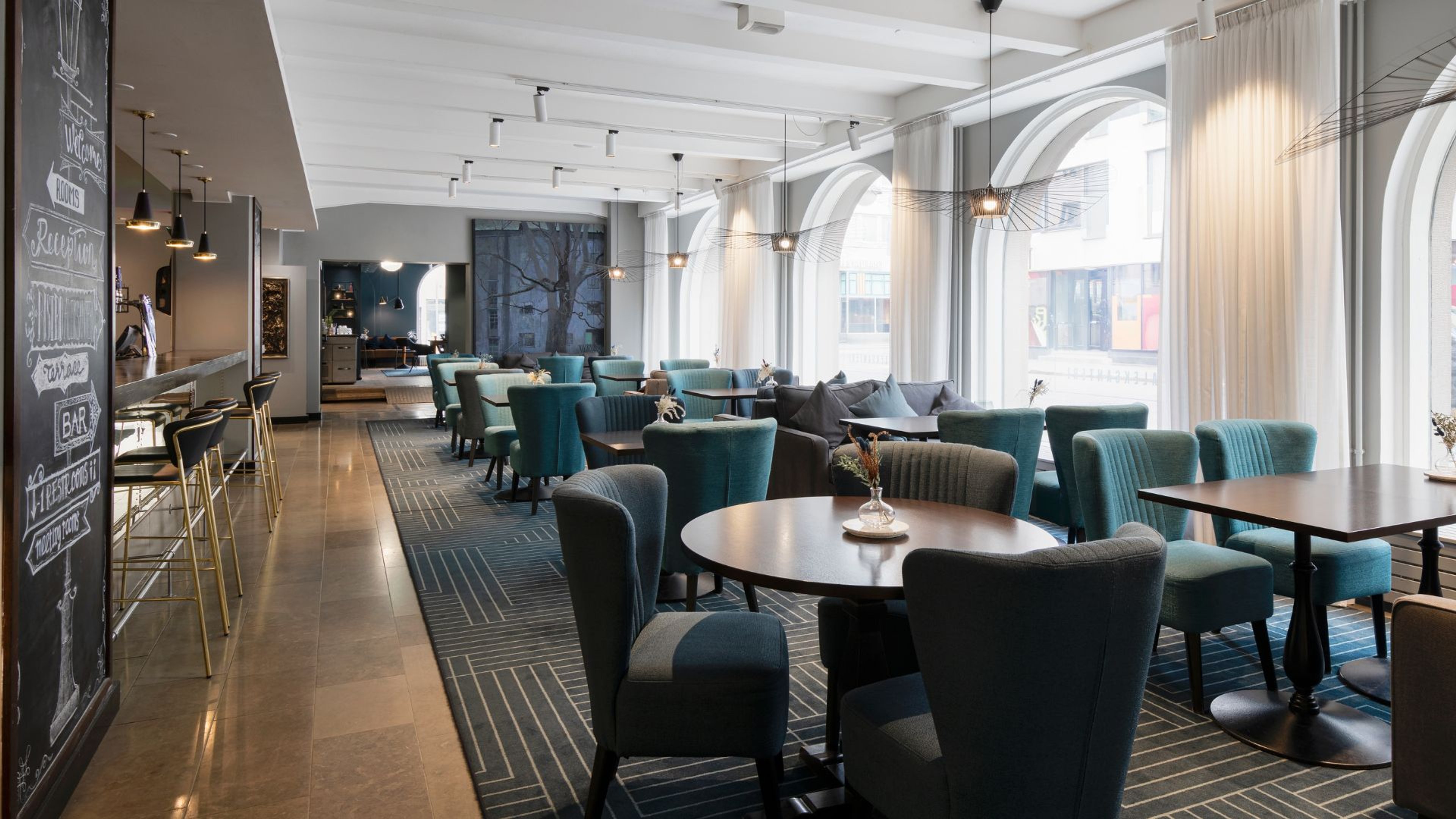 Moderni ravintola tai kahvila, jossa turkoosit nojatuolit, pyöreät pöydät, baaritiski vasemmalla ja suuret kaari-ikkunat. Tasainen lattia ja avara tila.
