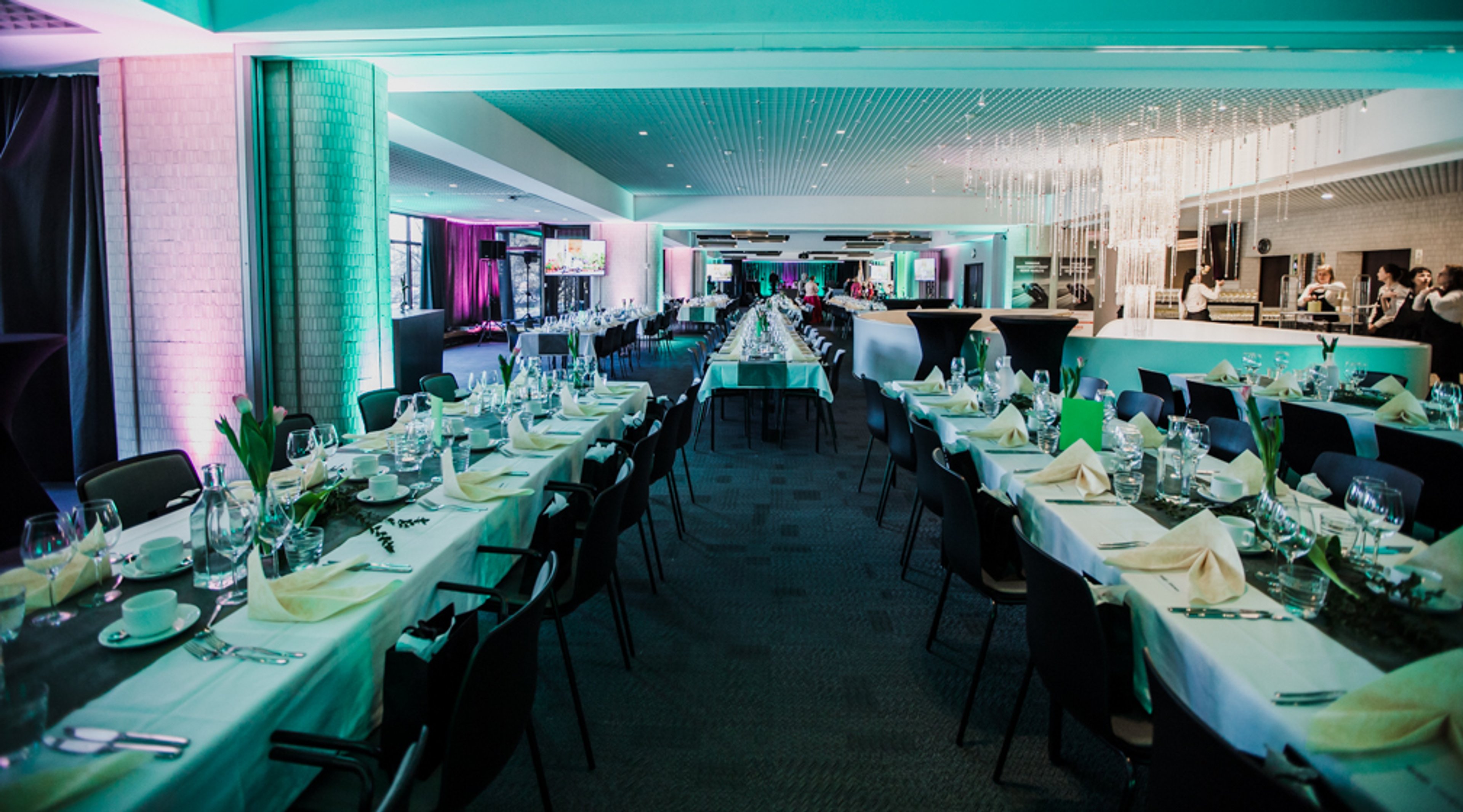 Juhlatila, jossa pitkät pöydät katettu turkoosilla ja mustalla väriteemalla, valaistus värillinen, tilava lattia-alue pöytien välissä.
