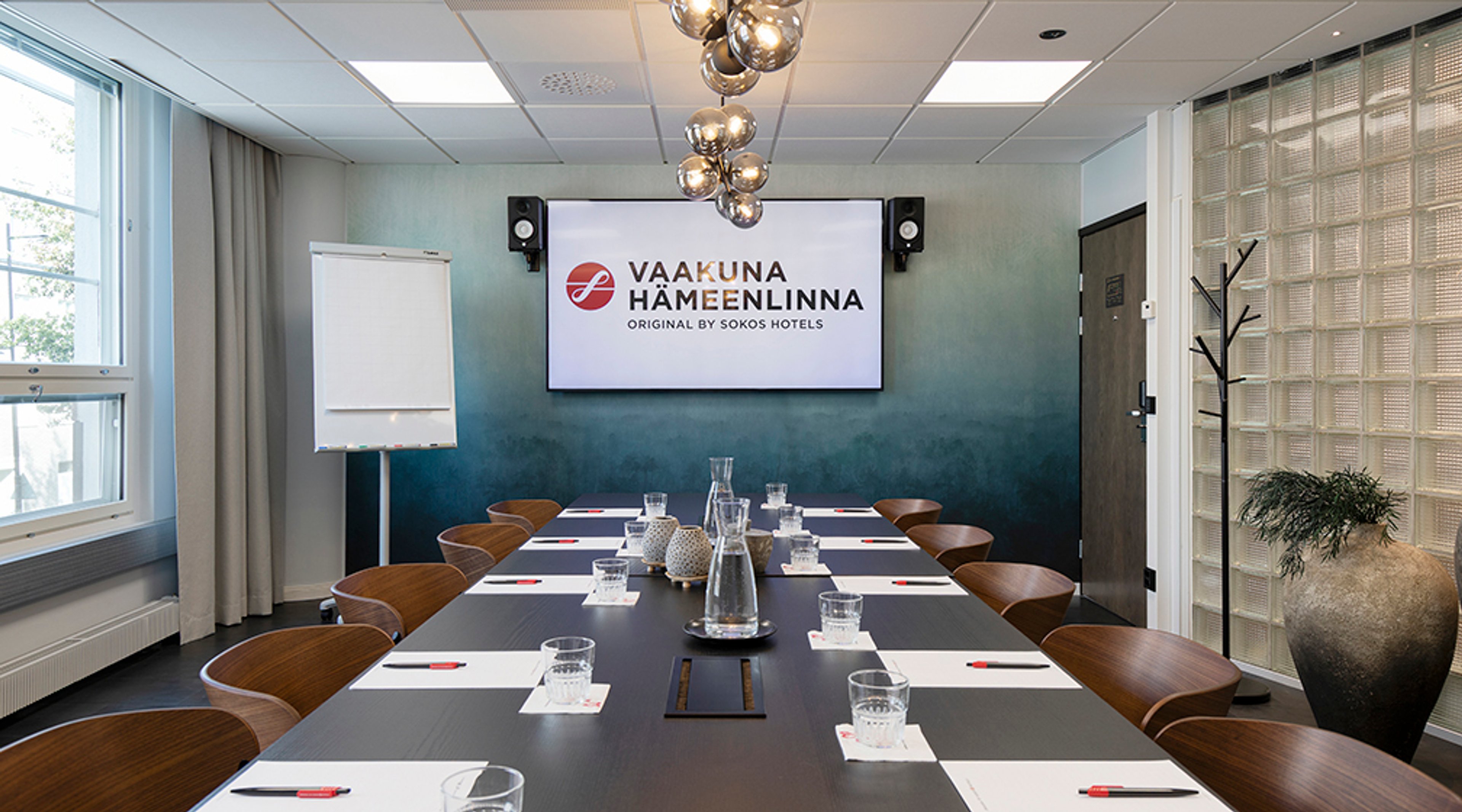 Moderni kokoushuone tummansinisellä seinällä ja suurella näytöllä. Pitkän pöydän ympärillä puisia tuoleja, pöydällä vesikannu ja lasit.
