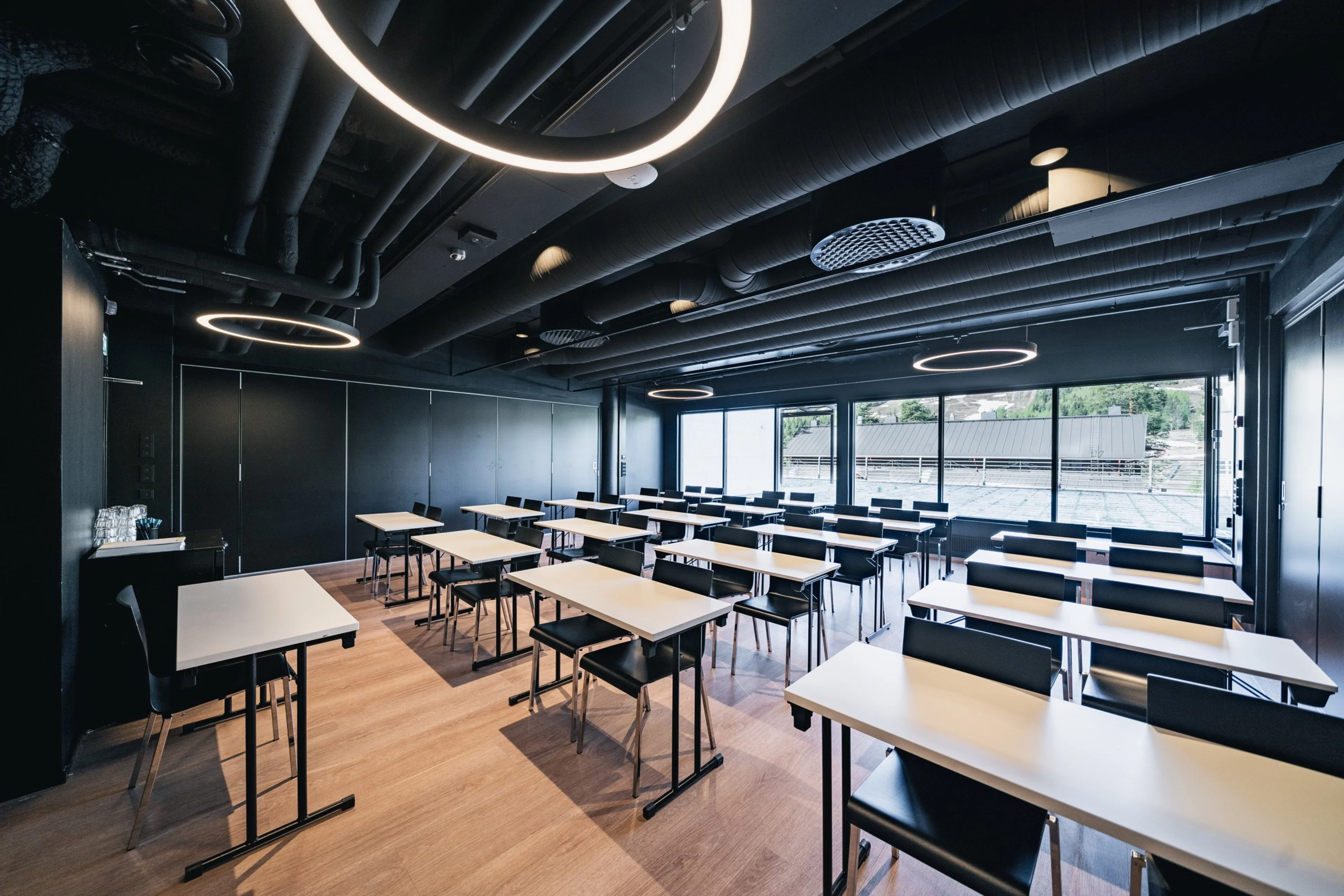 Moderni luokkahuone mustilla seinillä ja puulattialla. Tilassa valkoiset pöydät, mustat tuolit ja pyöreät kattovalaisimet. Suuret ikkunat tuovat luonnonvaloa.