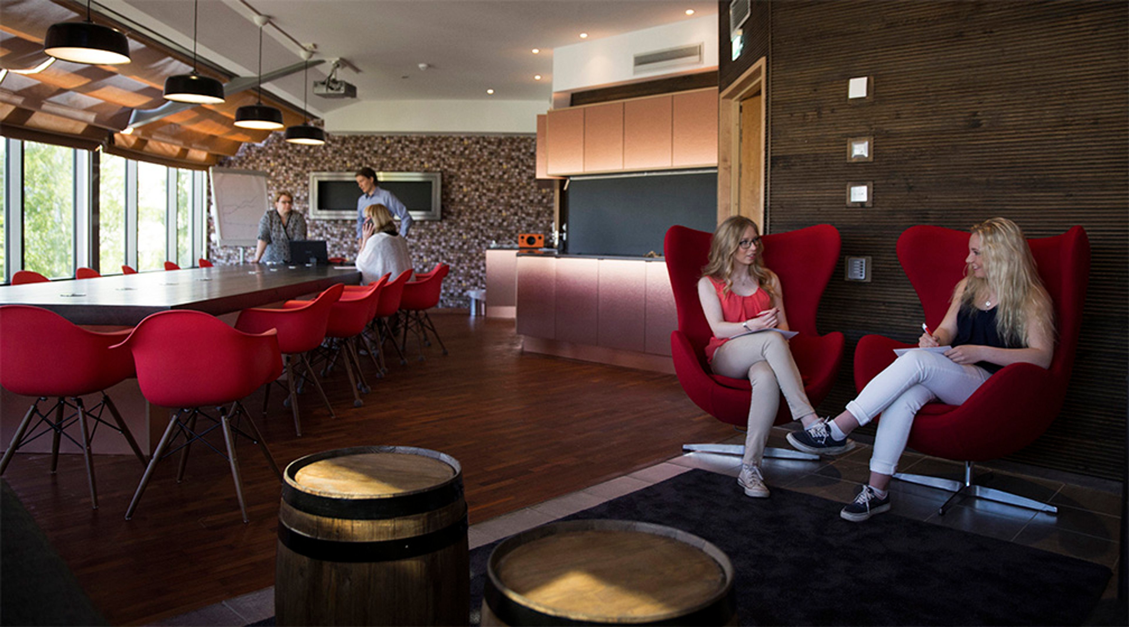 Moderni taukotila, jossa punaisia tuoleja, puulattia ja kaksi tynnyripöytää. Henkilöitä keskustelemassa avoimessa tilassa, jossa suuri pöytä ja keittiö.