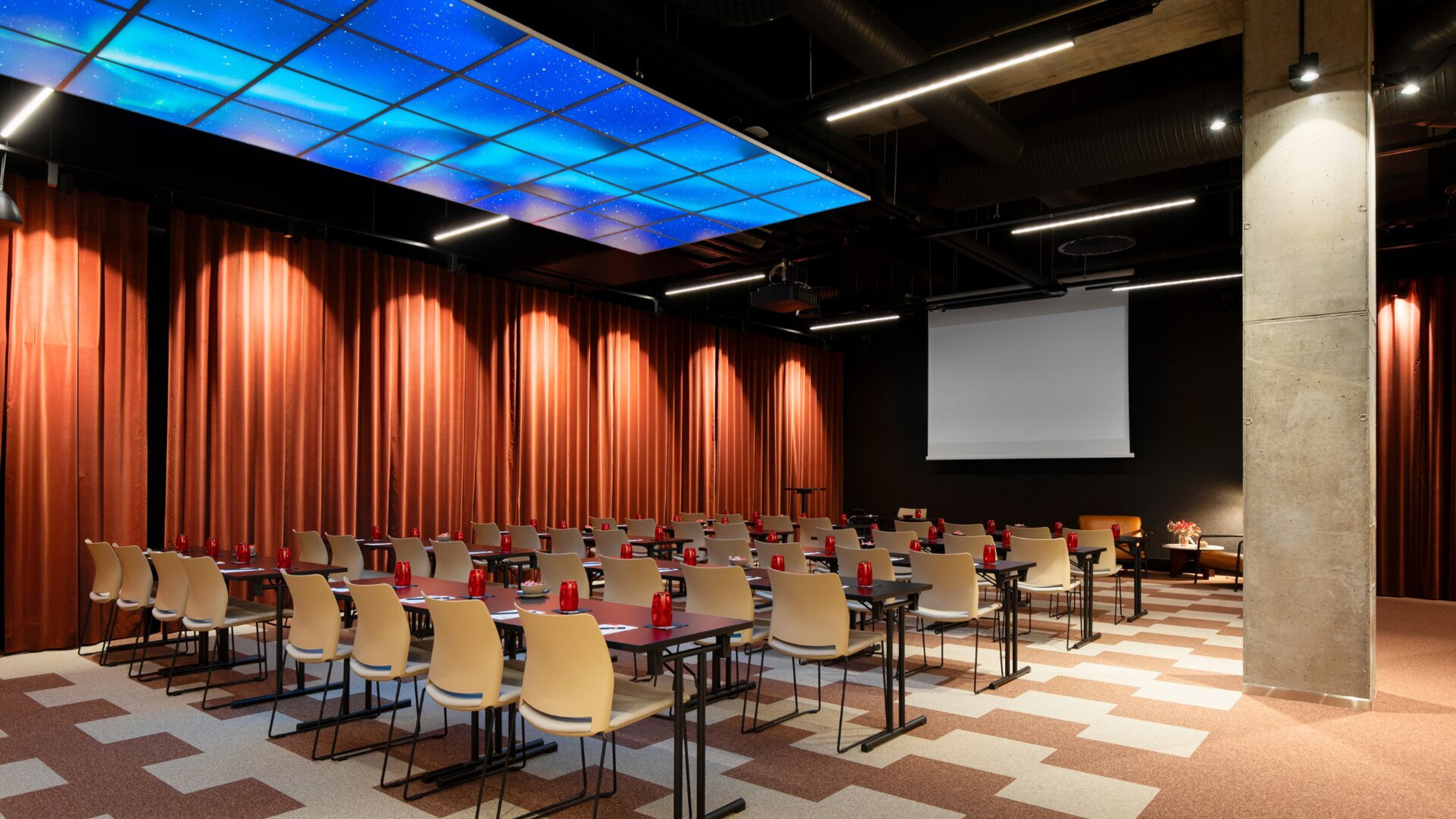 Moderni auditorio punaisilla verhoilla, valkoisilla tuoleilla ja tähtitaivasta kuvaavalla sinisellä katolla. Lattialla geometrinen kuviointi ja valkokangas edessä.