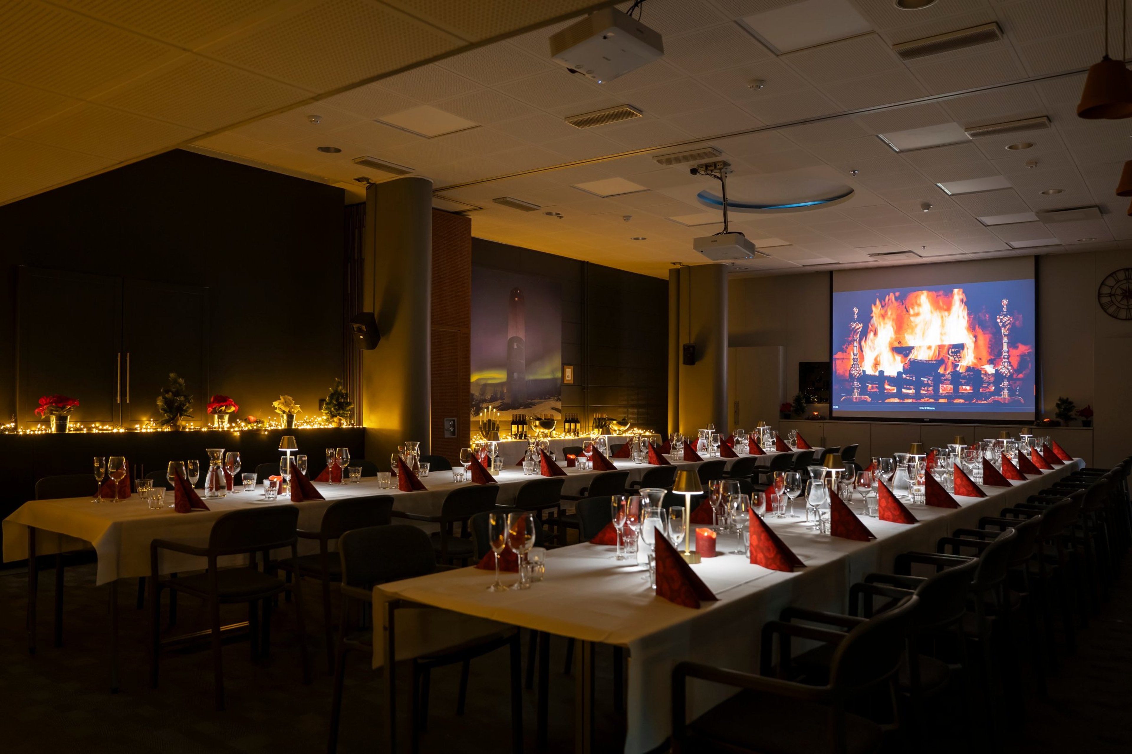 Juhlatila, jossa pöydät on katettu valkoisilla liinoilla ja punaisilla lautasliinoilla. Näytöllä näkyy takkatuli. Huoneessa on hämärä tunnelmavalaistus.